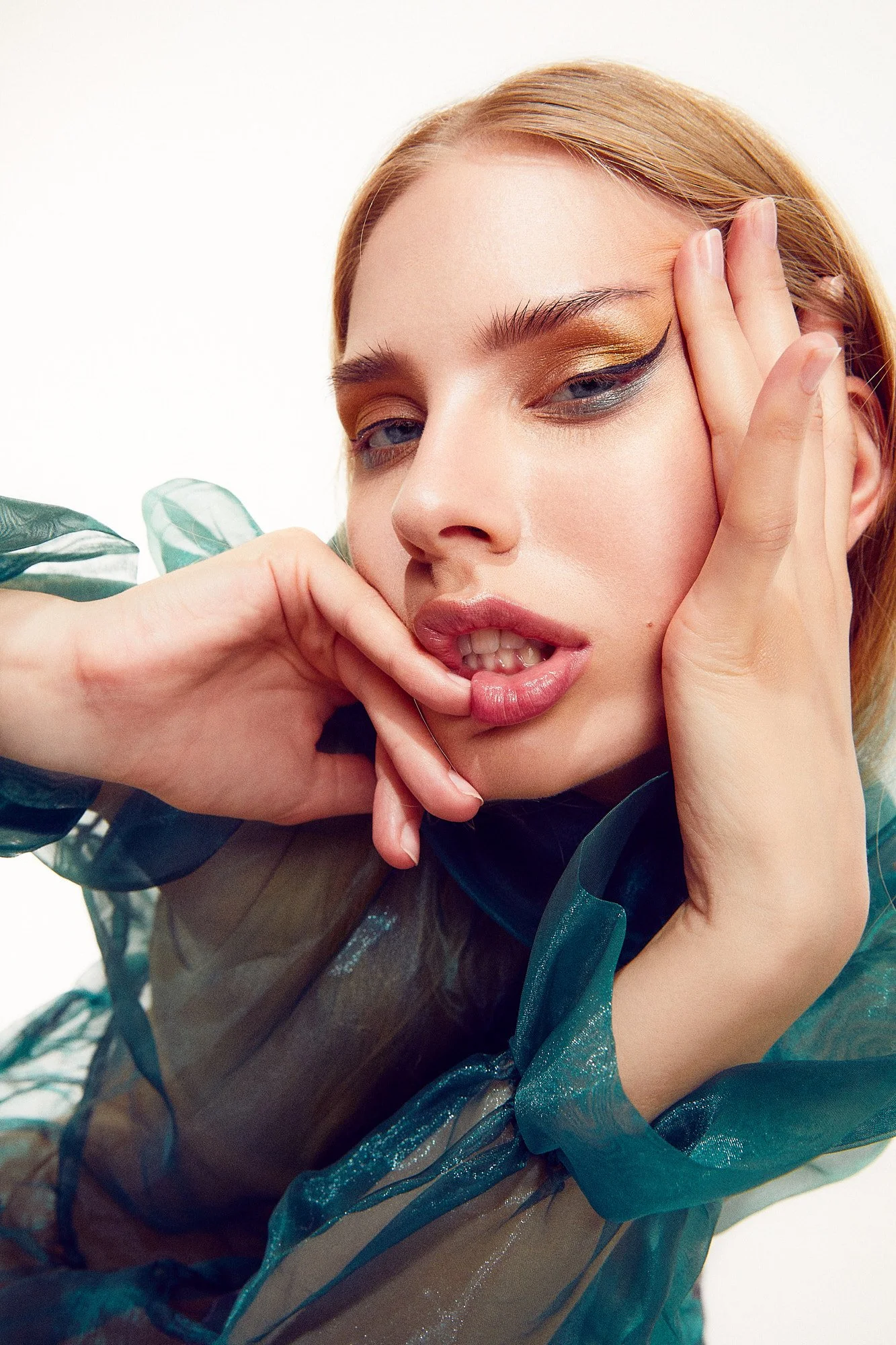 Close-up of a woman with red hair, bold eye makeup, and glossy lips, posing with her hand near her face.