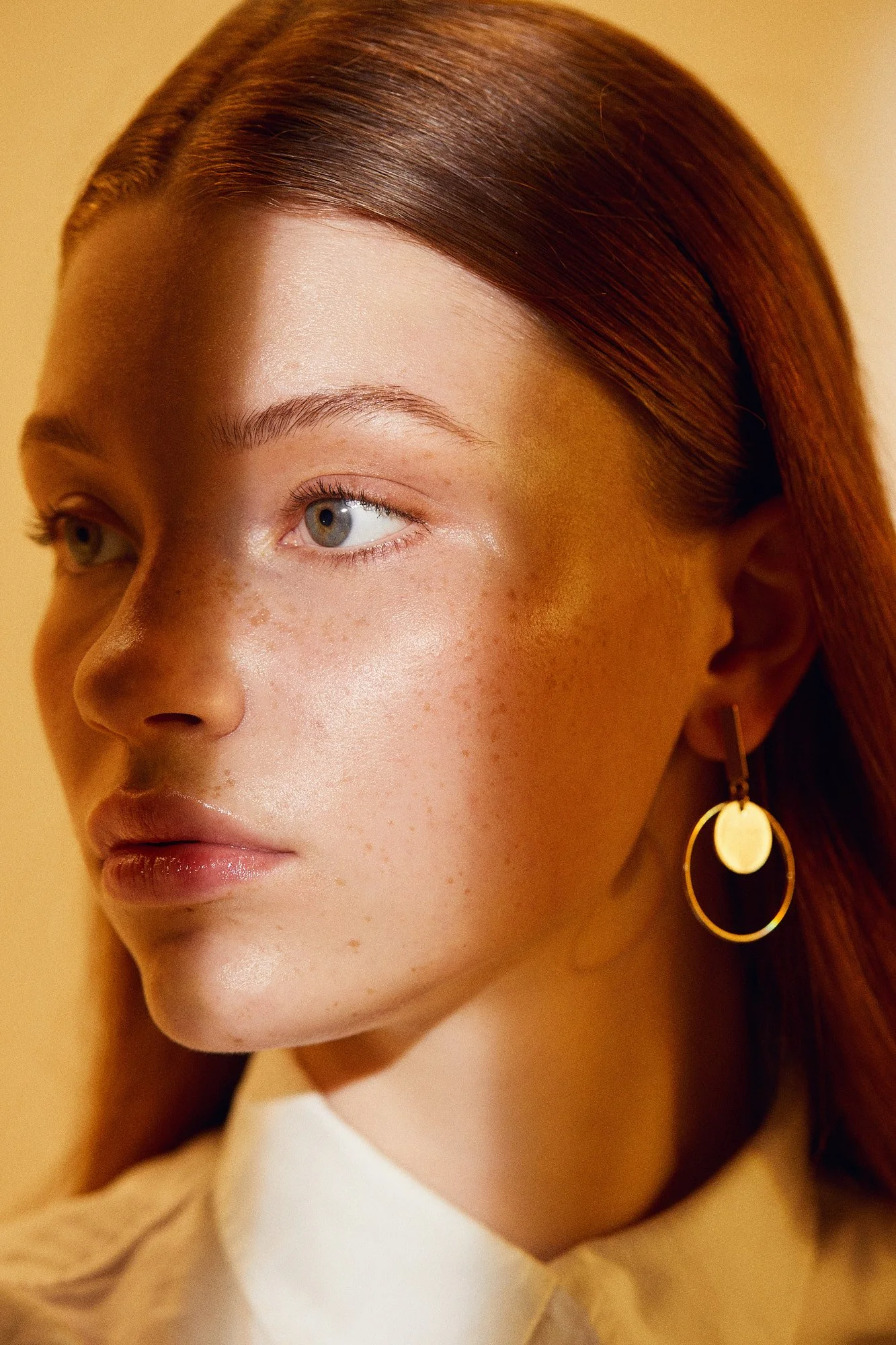 Close-up of a woman's face with red hair, tan skin, and gold hoop earring, gazing to the left, set against a warm yellow background.