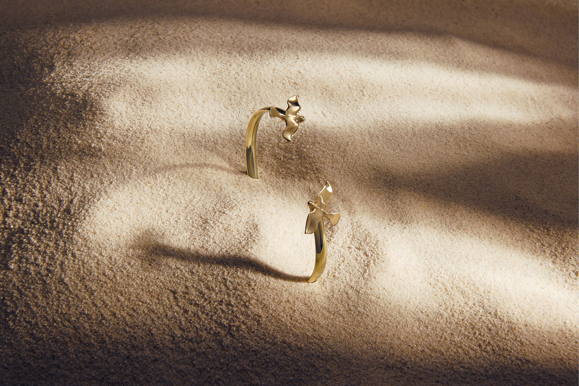 A close-up view of a gold ring with bow-shaped designs, partially embedded in fine, light brown sand, with gentle shadows and a warm, soft lighting effect.