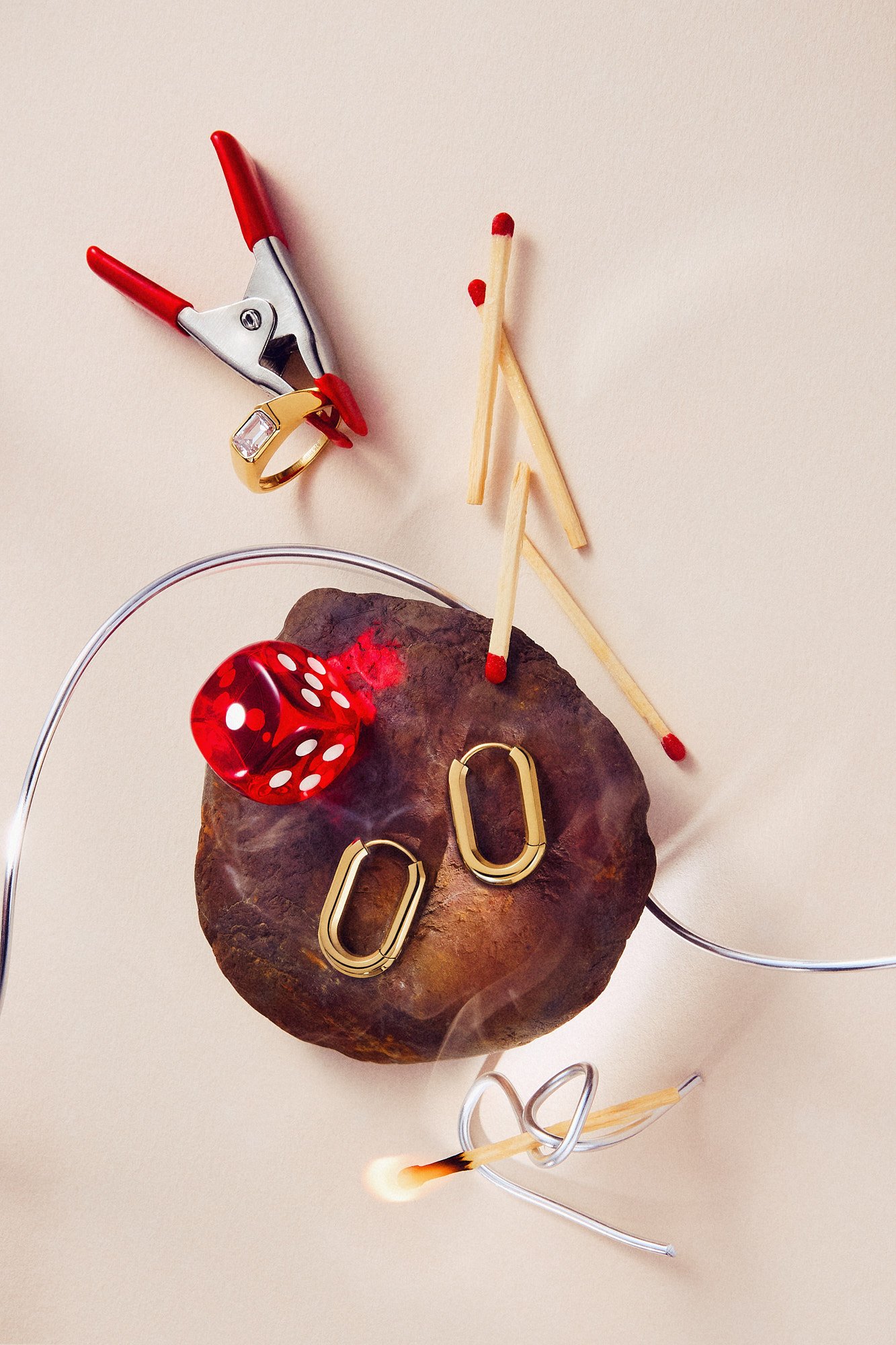 A rock with red dice-shaped beads and gold paperclip jewelry, a red ring with a clear gemstone, decorative wire, and wooden matches with red tips on a light background.