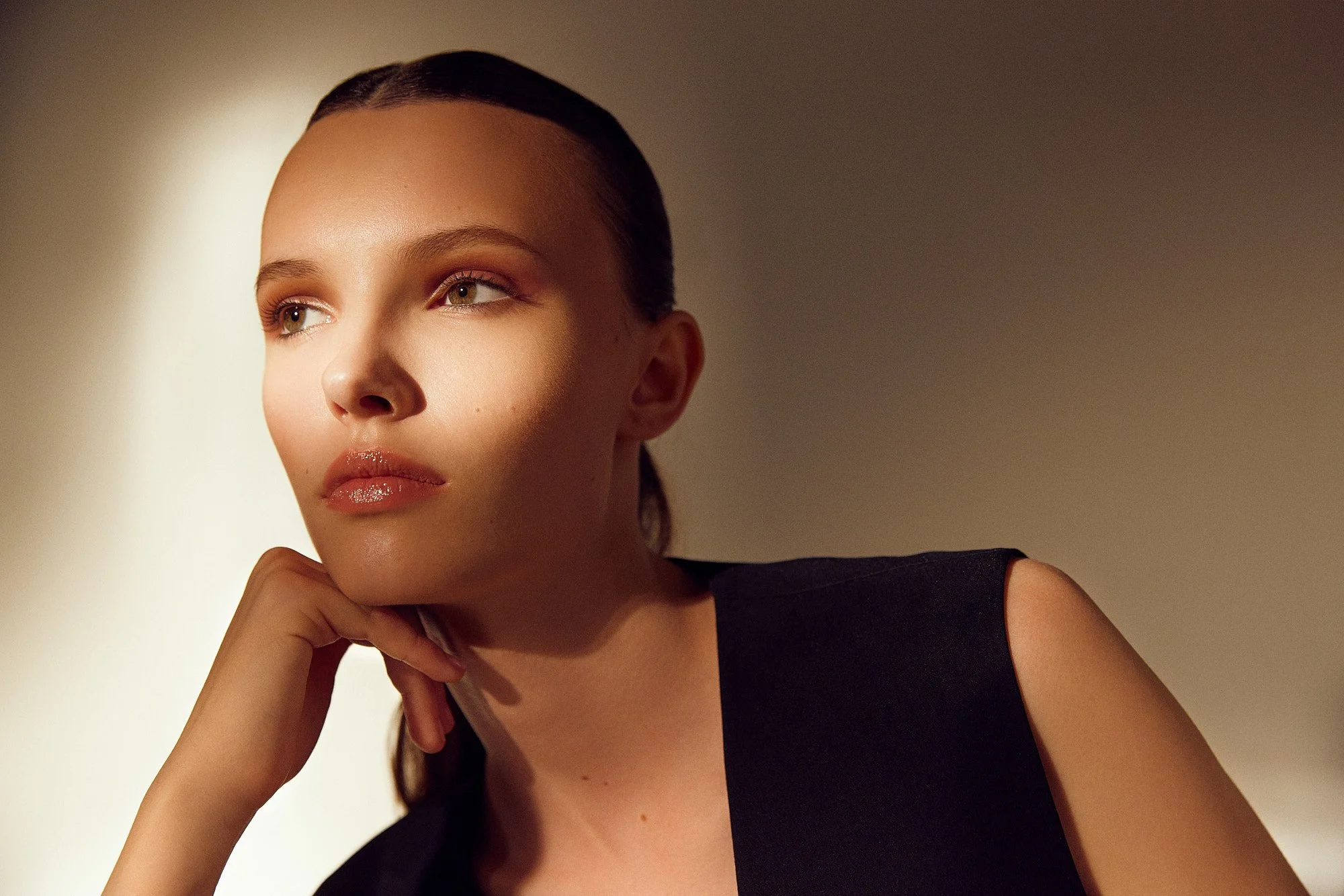 Portrait of a woman with fair skin and light eye color, resting her chin on her hand, with dark hair tied back, wearing a black sleeveless top, in front of a plain beige background.