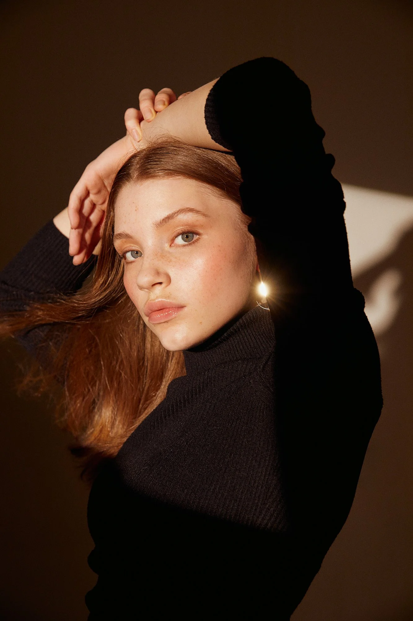 Portrait of a young woman with long, wavy auburn hair, light skin, and freckles, wearing a black long-sleeve top, with her left arm raised and hand resting on her head, against a dark background.