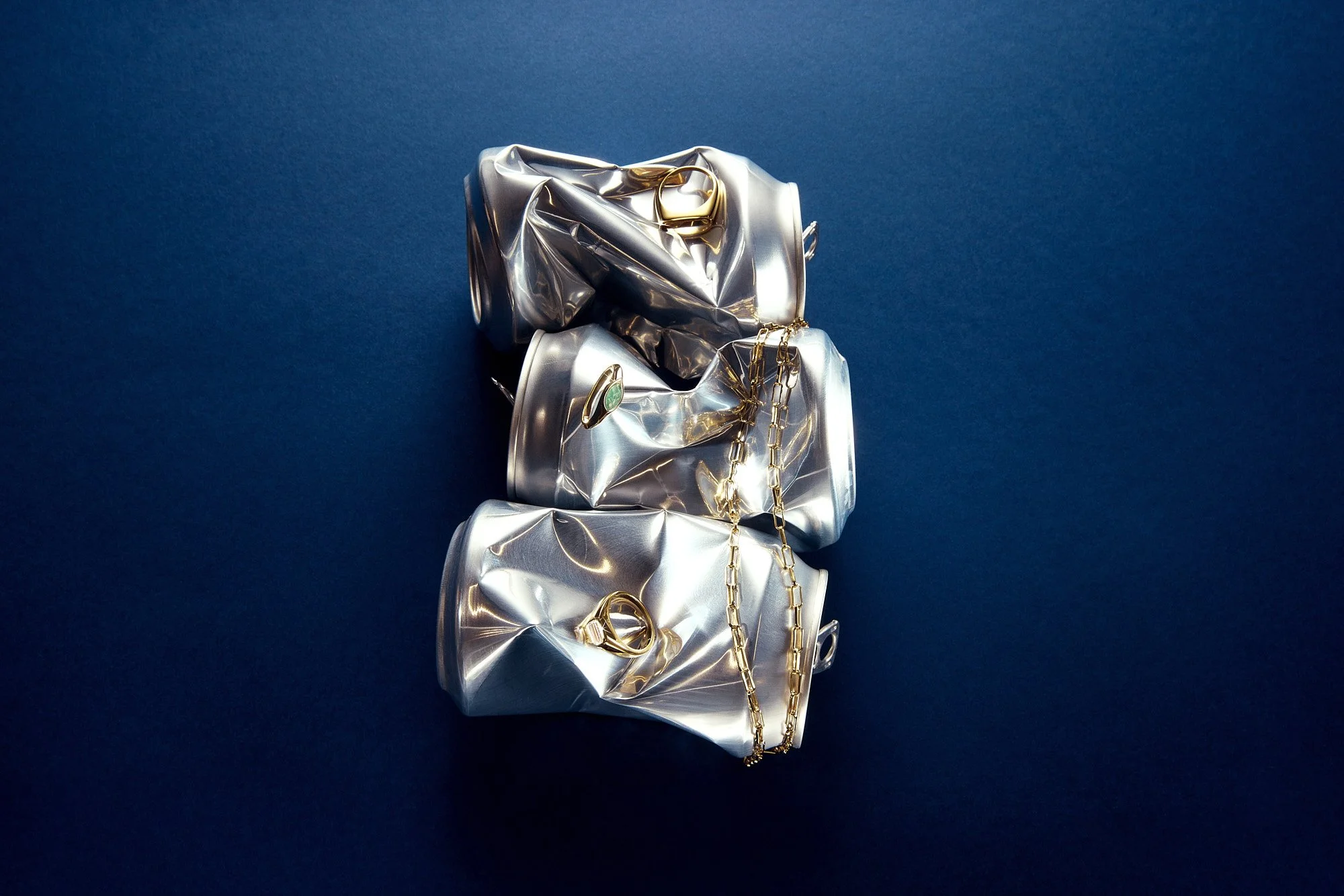 Crushed aluminum cans with gold pdpaola jewelry on a dark blue background.
