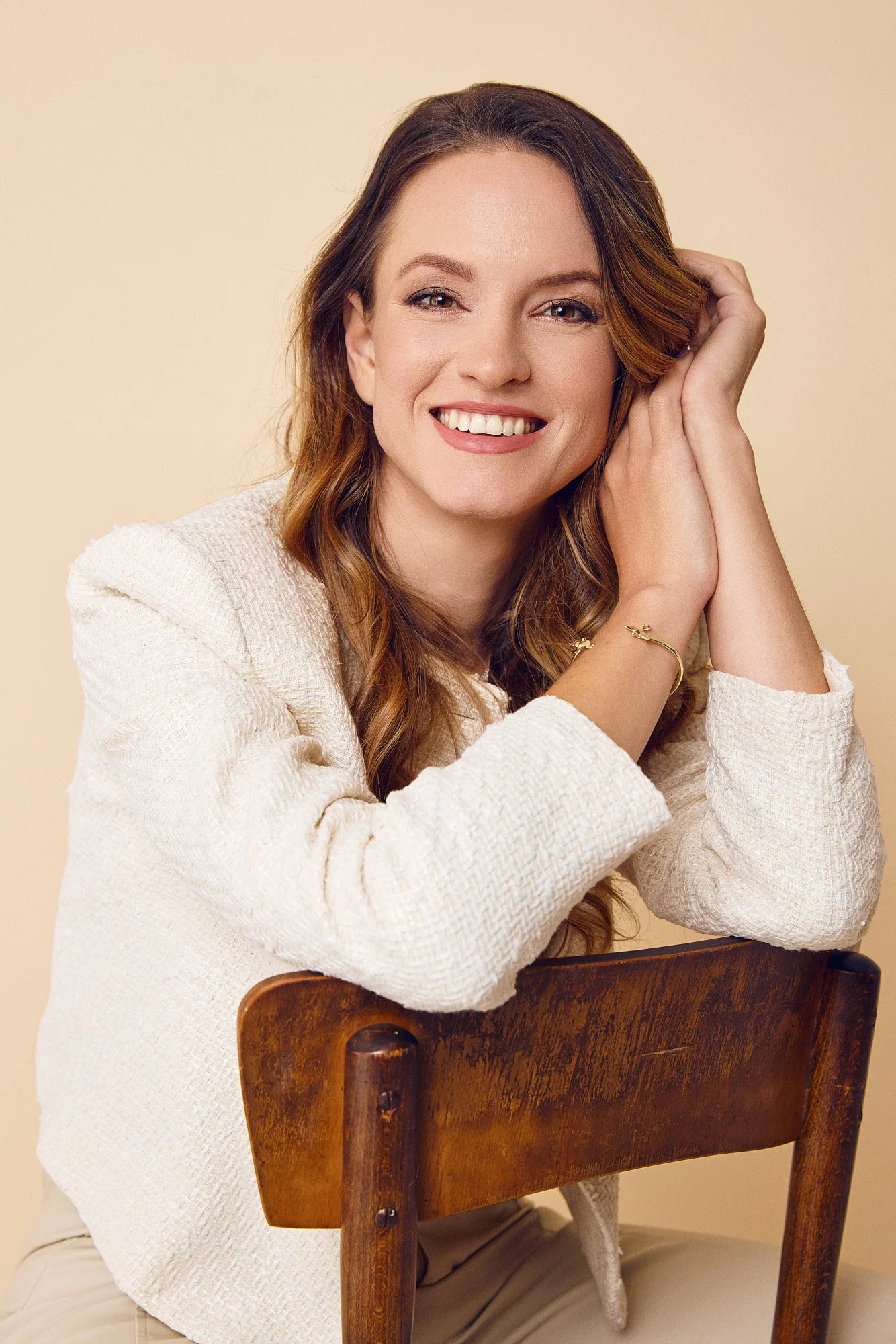 A woman with long brown hair, smiling, wearing a cream-colored textured sweater, seated behind a wooden chair with her arms resting on its back, posing against a plain beige background.