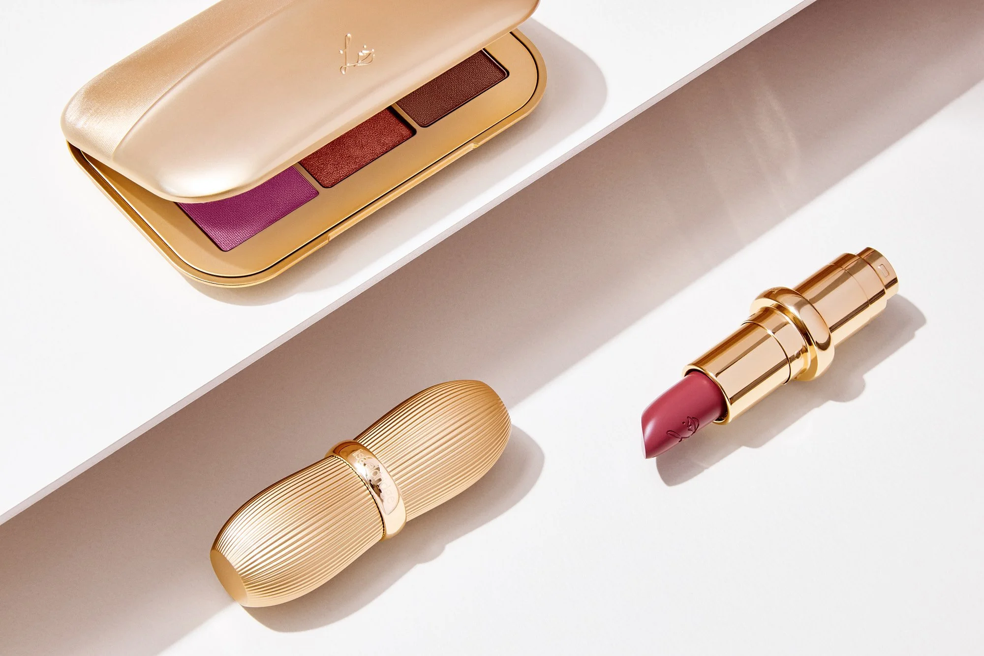 Close-up of a beige Lisa Eldridge makeup palette with three eyeshadows in purple, red, and brown, along with a pink lipstick and two gold-colored lipstick tubes on white background.