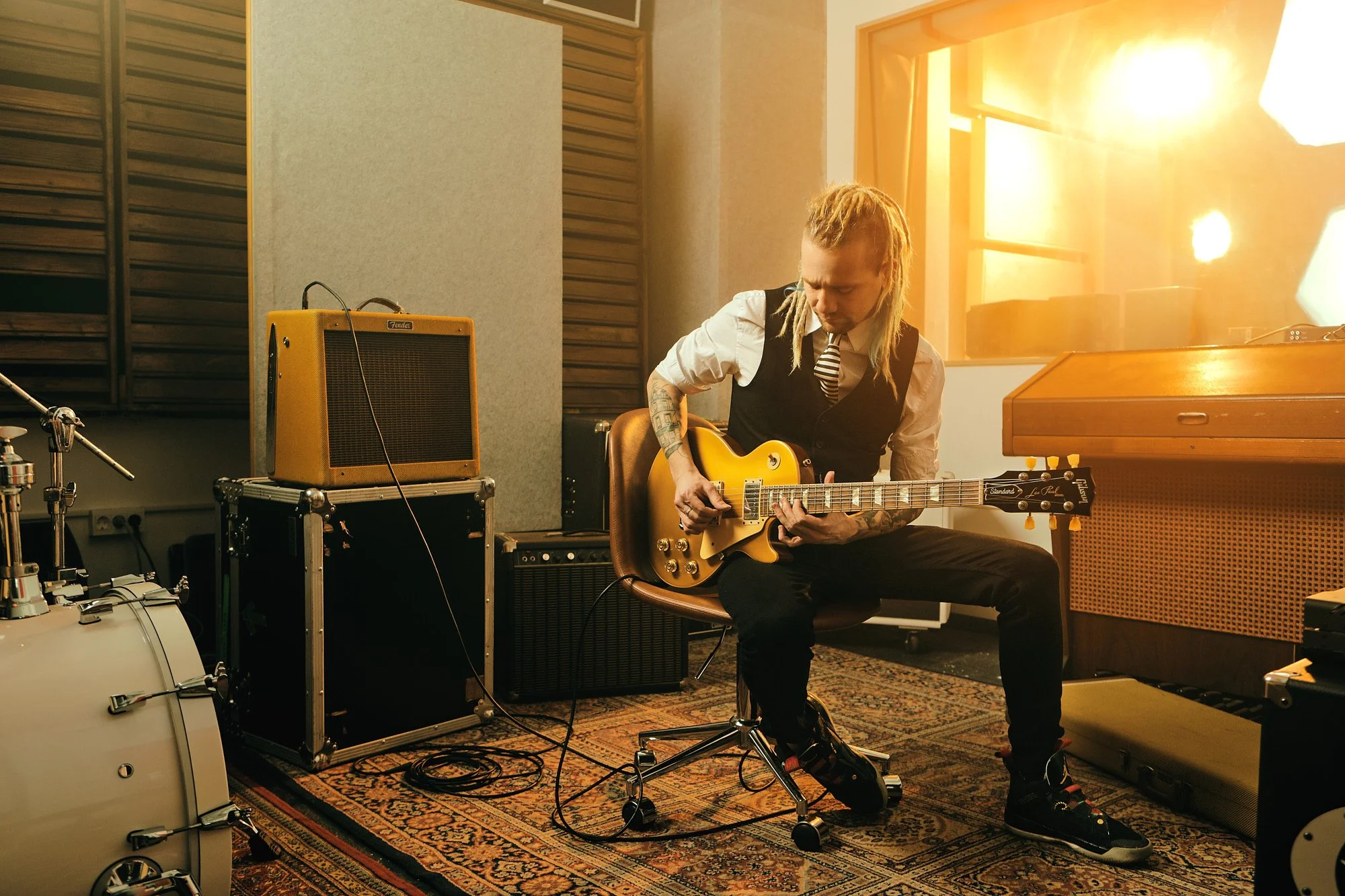 A musician with dreadlocks playing a yellow electric guitar in a recording studio, surrounded by audio equipment and a drum set, with warm sunlight coming through a window.