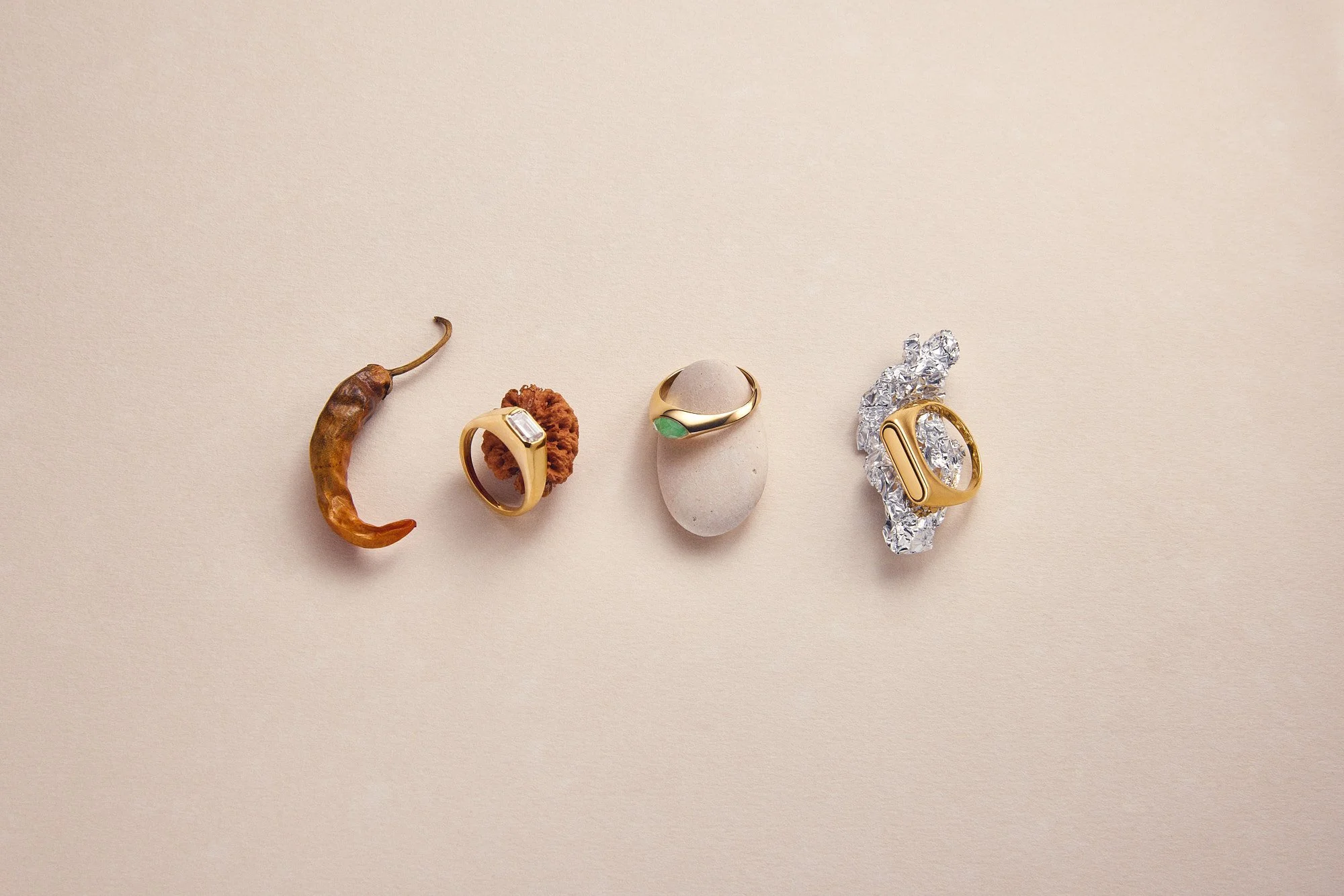 A collection of pdpaola jewelry and a dried chili pepper arranged on a plain background, including rings, a brooch, and a stone.