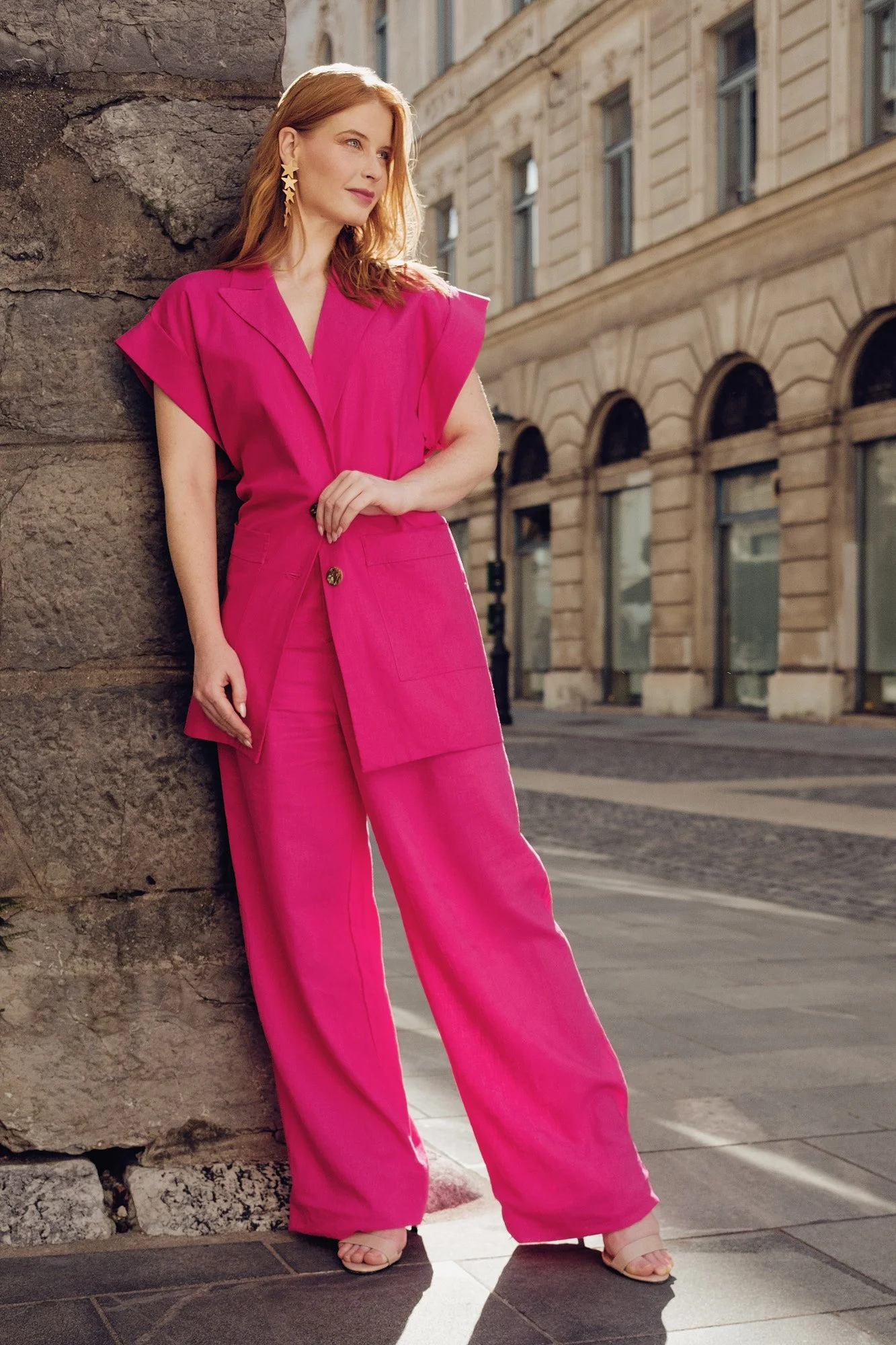 A woman with red hair wearing a bright pink pantsuit and star-shaped earrings, standing against a stone wall on a city street, with historic buildings in the background.