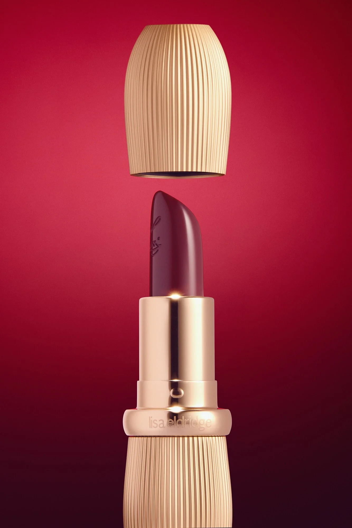 Close-up of a Lisa Eldridge lipstick tube with a carved face on the lipstick, set against a red background. The lipstick is in a beige and metallic casing with a ribbed design. The brand name, Lisa Aldridge, is engraved on the casing.