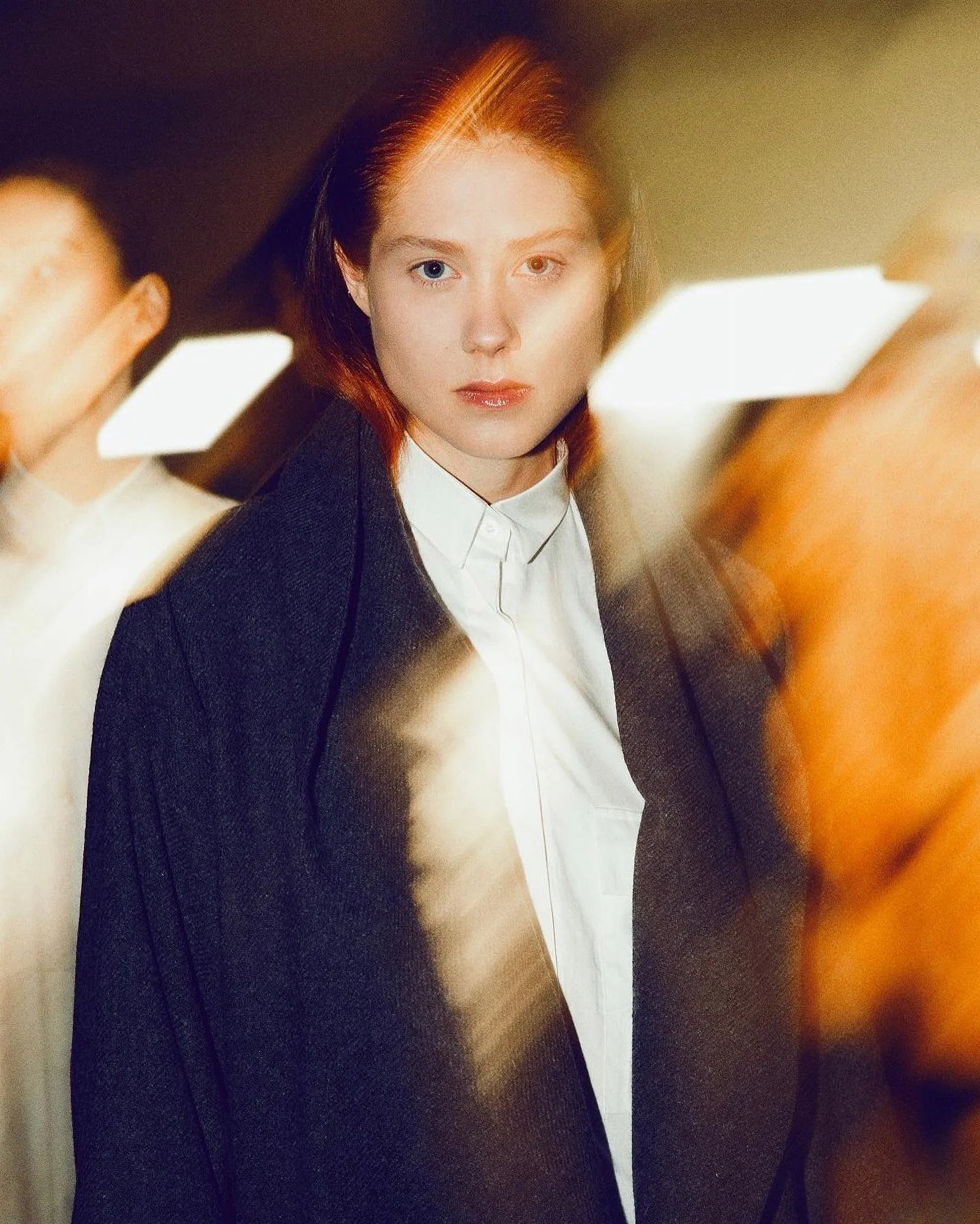 A woman with red hair and heterochromatic eyes (one blue and one hazel) is looking directly at the camera; she is dressed in a white shirt and black blazer, with a serious expression.
