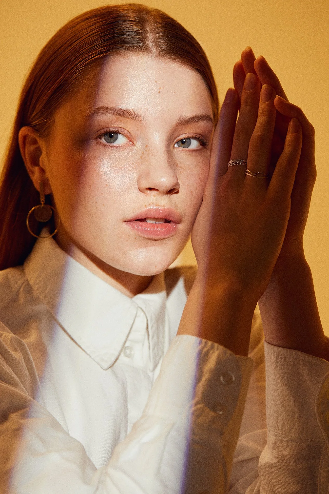 Close-up of a woman with red hair, fair skin, blue eyes, freckles, wearing a white collared shirt and gold jewelry, holding her hands near her face against a yellow background.