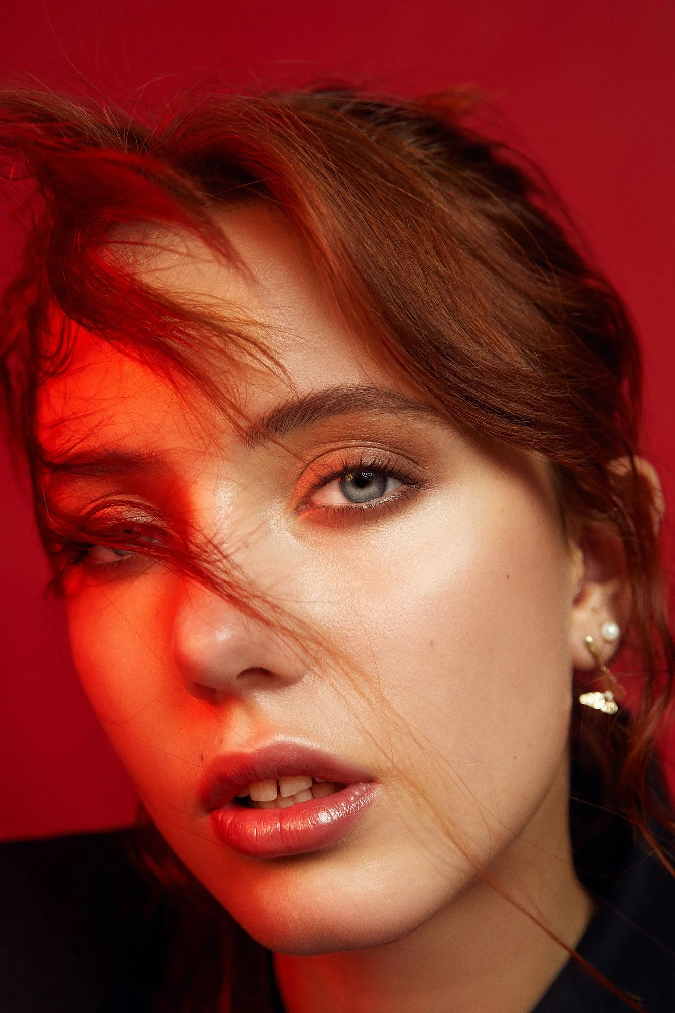 Close-up of a woman with red hair, blue eyes, makeup, and pearl earrings, set against a red background.
