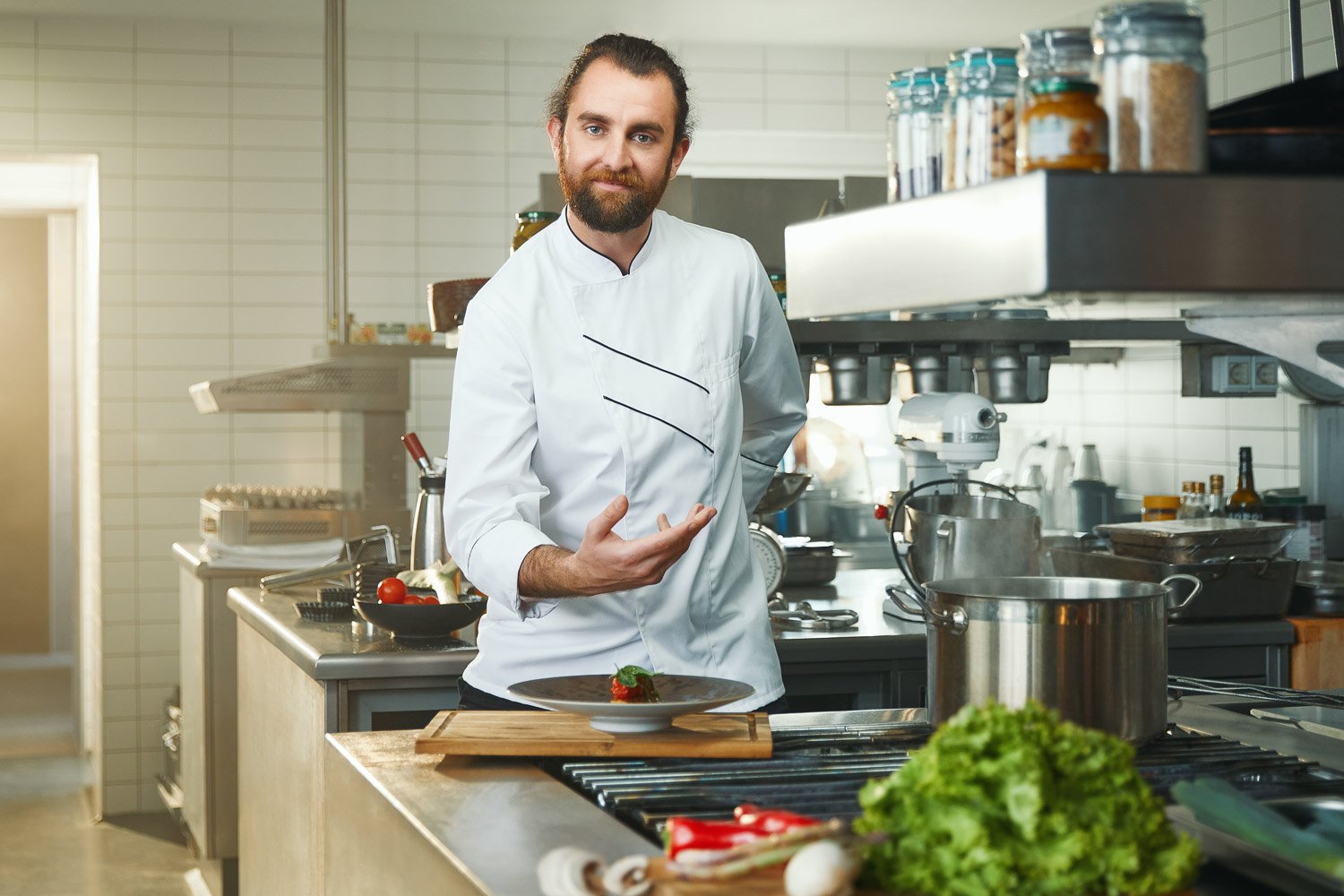 A male chef in a kitchen cooking, with a plate of food in front of him and kitchen utensils and ingredients around.