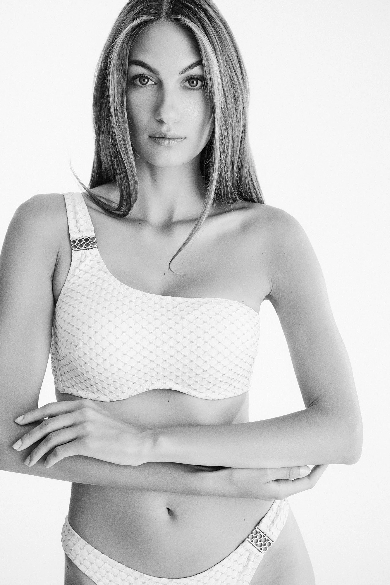Black and white portrait of a woman with long hair, wearing a textured crop top and matching bottoms, standing with her arms crossed, against a plain background.
