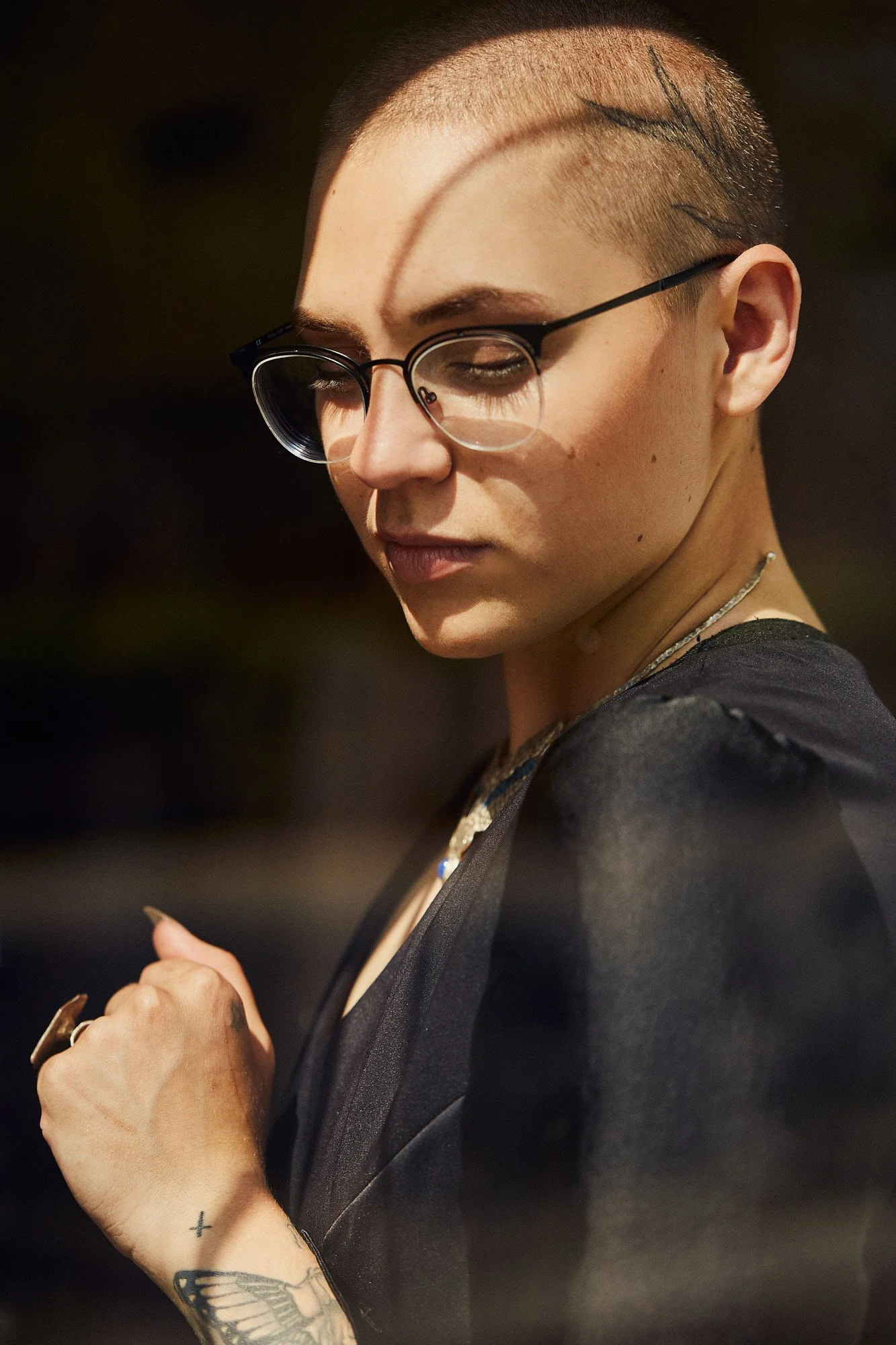 A person with short, shaved hair and tattoos on their arm and face, wearing glasses, is looking down at their phone and holding it with both hands.