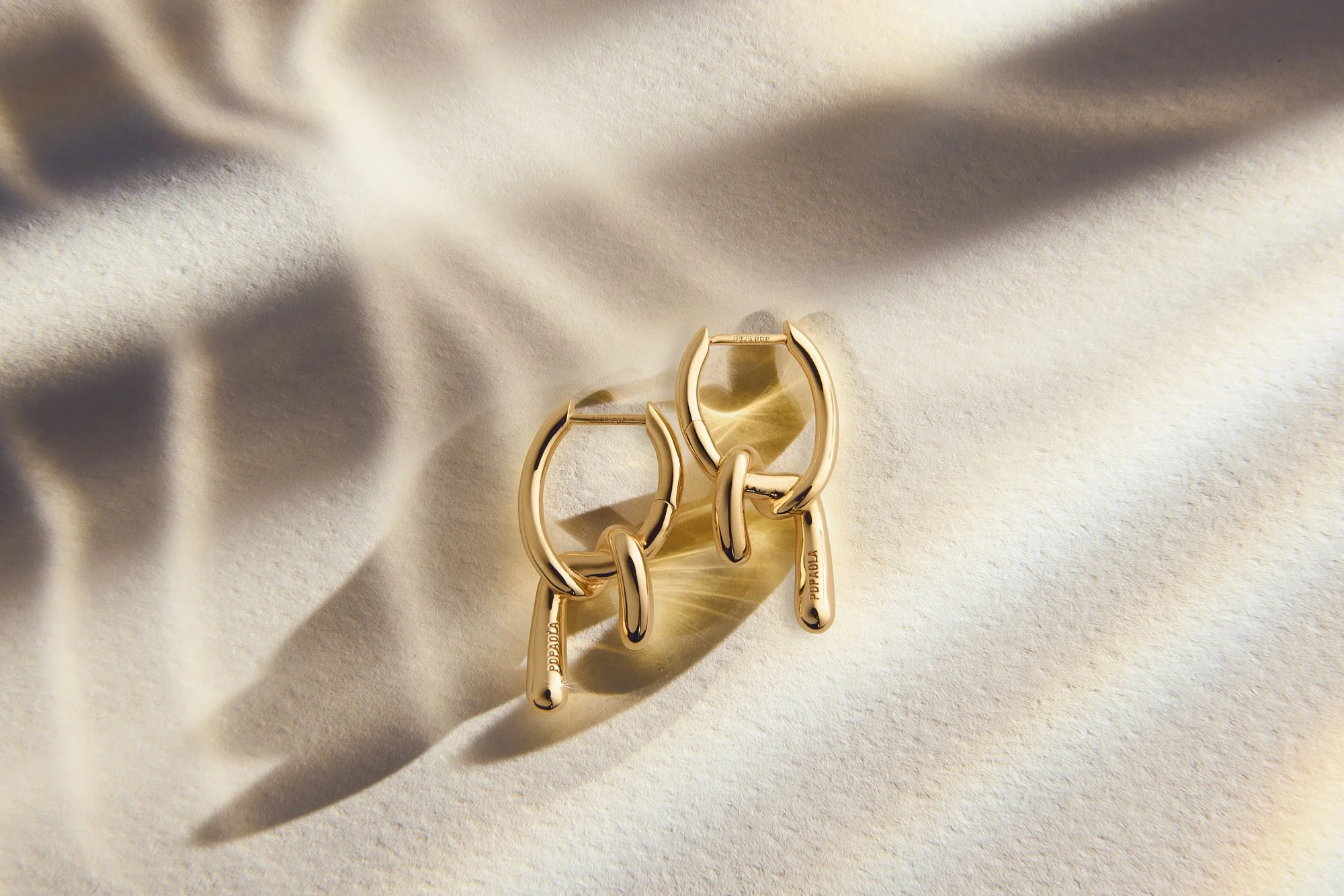Gold hoop earrings with dangling tag that reads 'POPPAJA' laid on a cream-colored fabric surface with soft shadows.