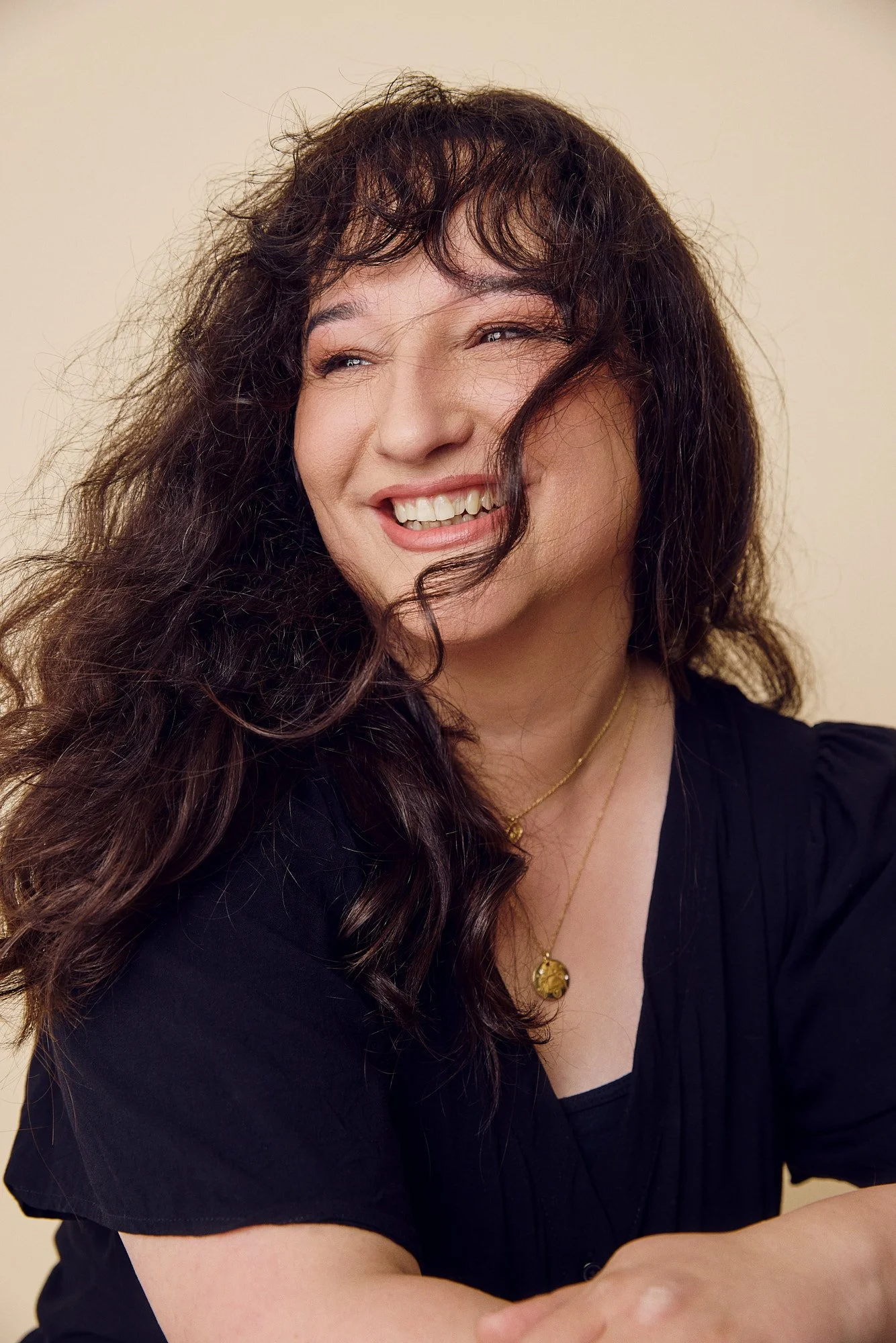 A woman with curly brown hair smiling and looking to the side, wearing a black top and gold Studio ORYN jewelry, standing against a plain background.