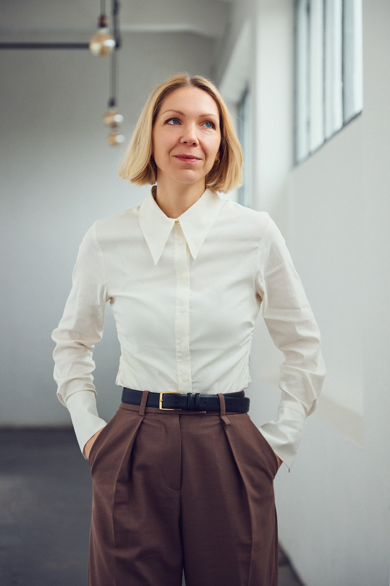 A woman with blond hair wearing a white button-up shirt and brown pants, standing indoors with her hands in her pockets.