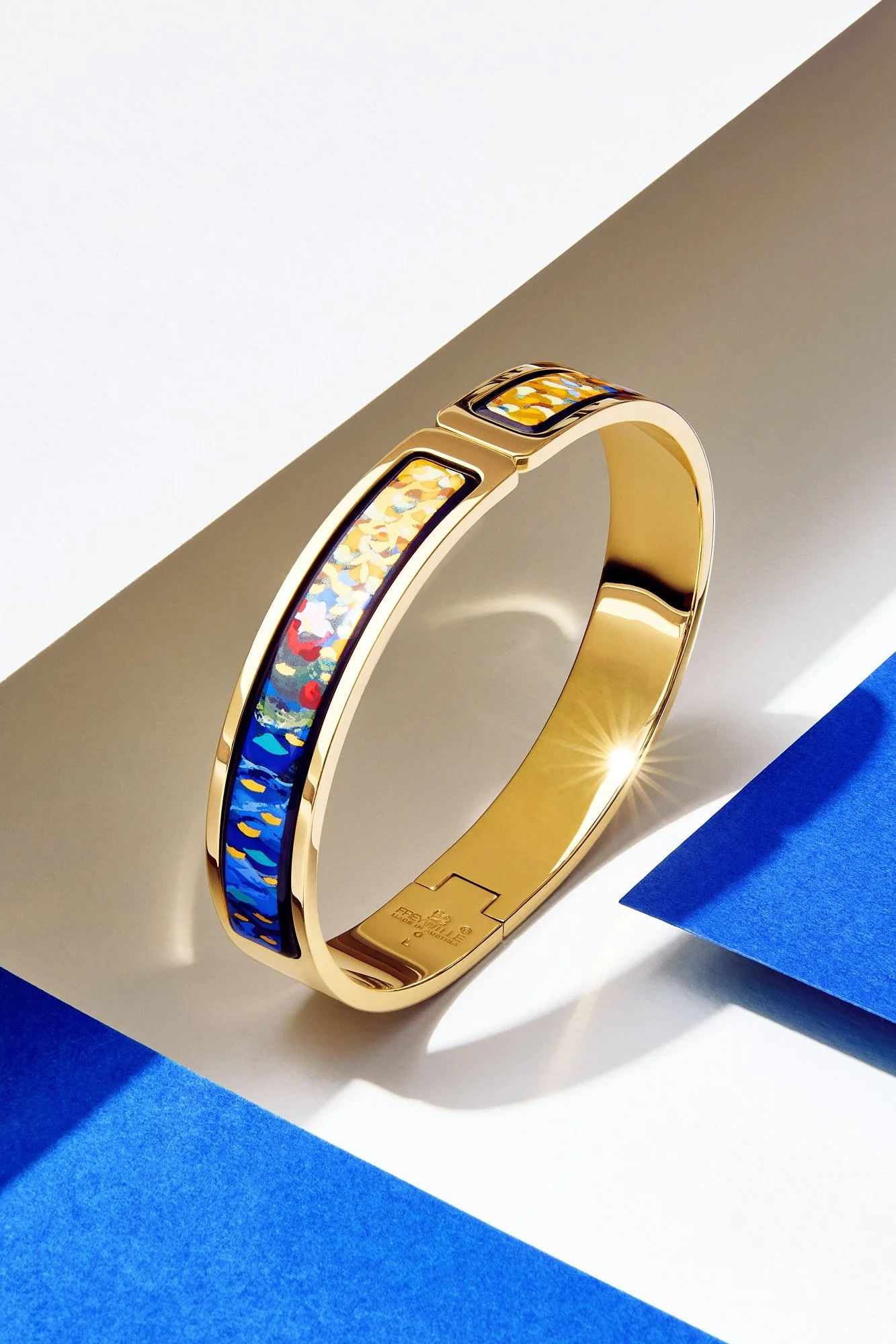 A gold Frey Wille bracelet with a colorful abstract pattern on the top, placed on a white and blue surface with a blue paper underneath.