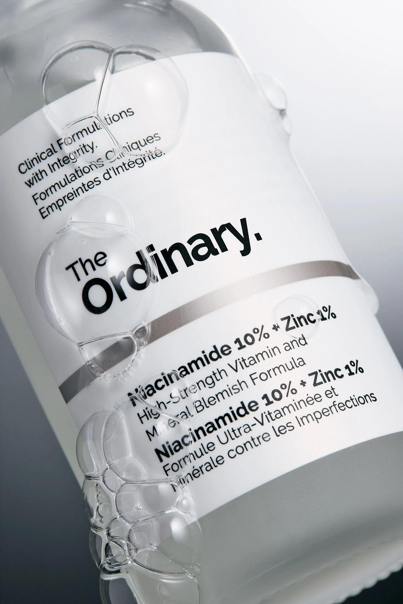 A bottle labeled 'The ordinary' with text about Niacinamide 10% + Zinc 1% and its benefits, with transparent glass capsules and bubbles surrounding it.