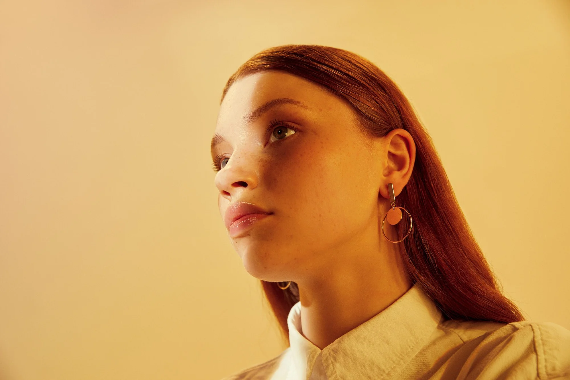 Close-up side profile of a young woman with red hair, wearing dangling earrings, against a plain yellow background.