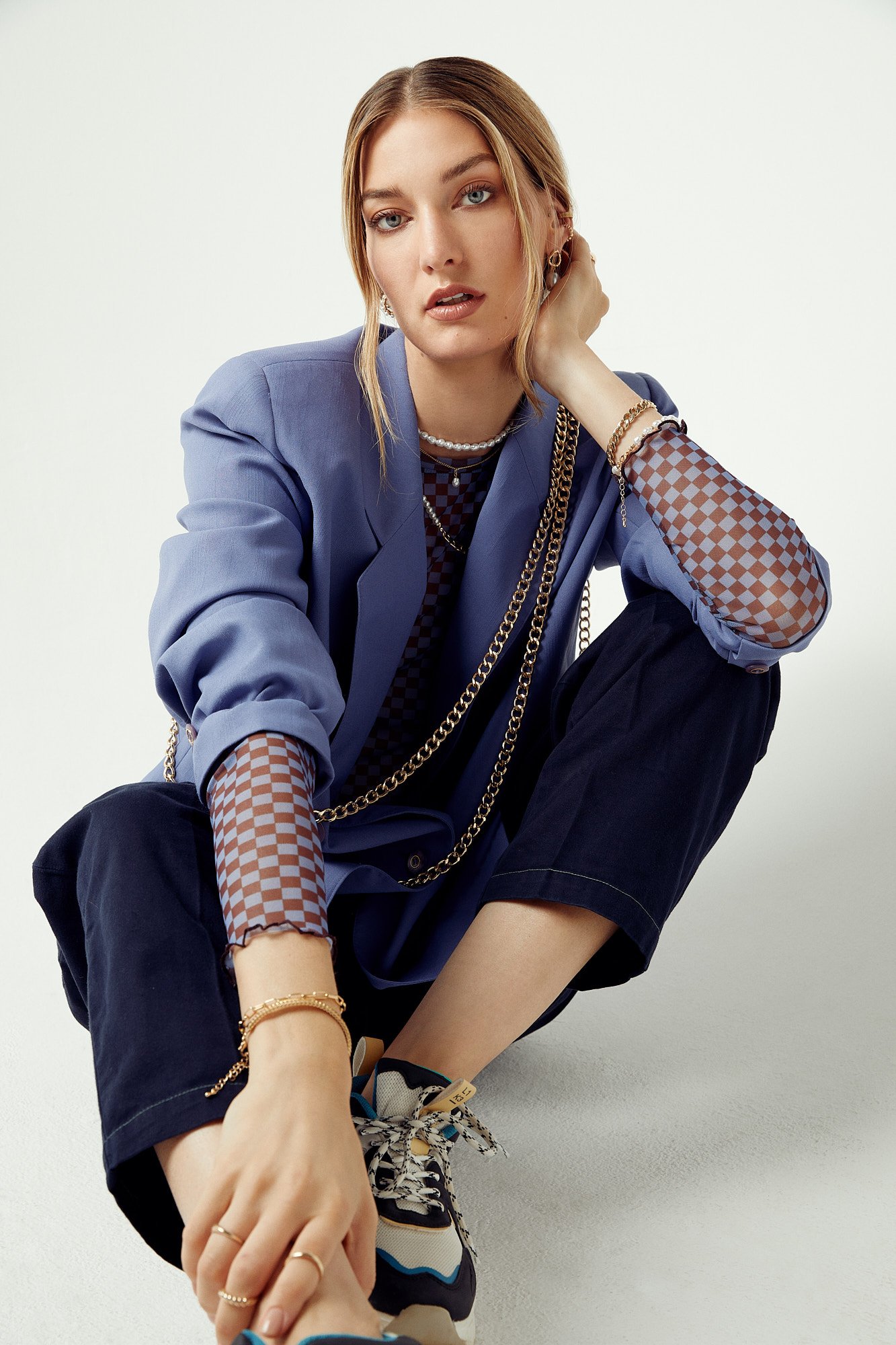 A young woman with  layered jewelry including necklaces, bracelets, and earrings and is gazing directly at the camera with a relaxed pose.