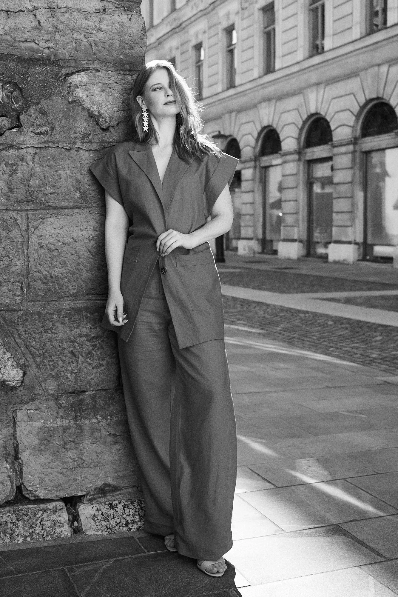 A woman with shoulder-length hair, wearing large star-shaped earrings, a sleeveless blazer, and wide-leg pants, leaning against a stone wall on a city street.