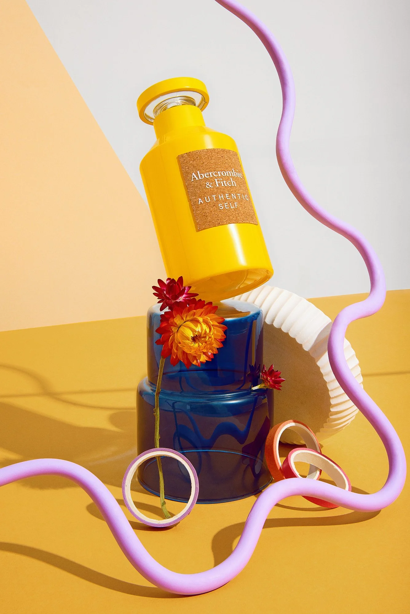 A yellow Abercrombie & Fitch soap pump bottle with a cork label that says 'Authentic Self' is placed on a blue glass vase, surrounded by colorful flowers, pink and purple curvy straws, and rings, with a yellow and pastel background.