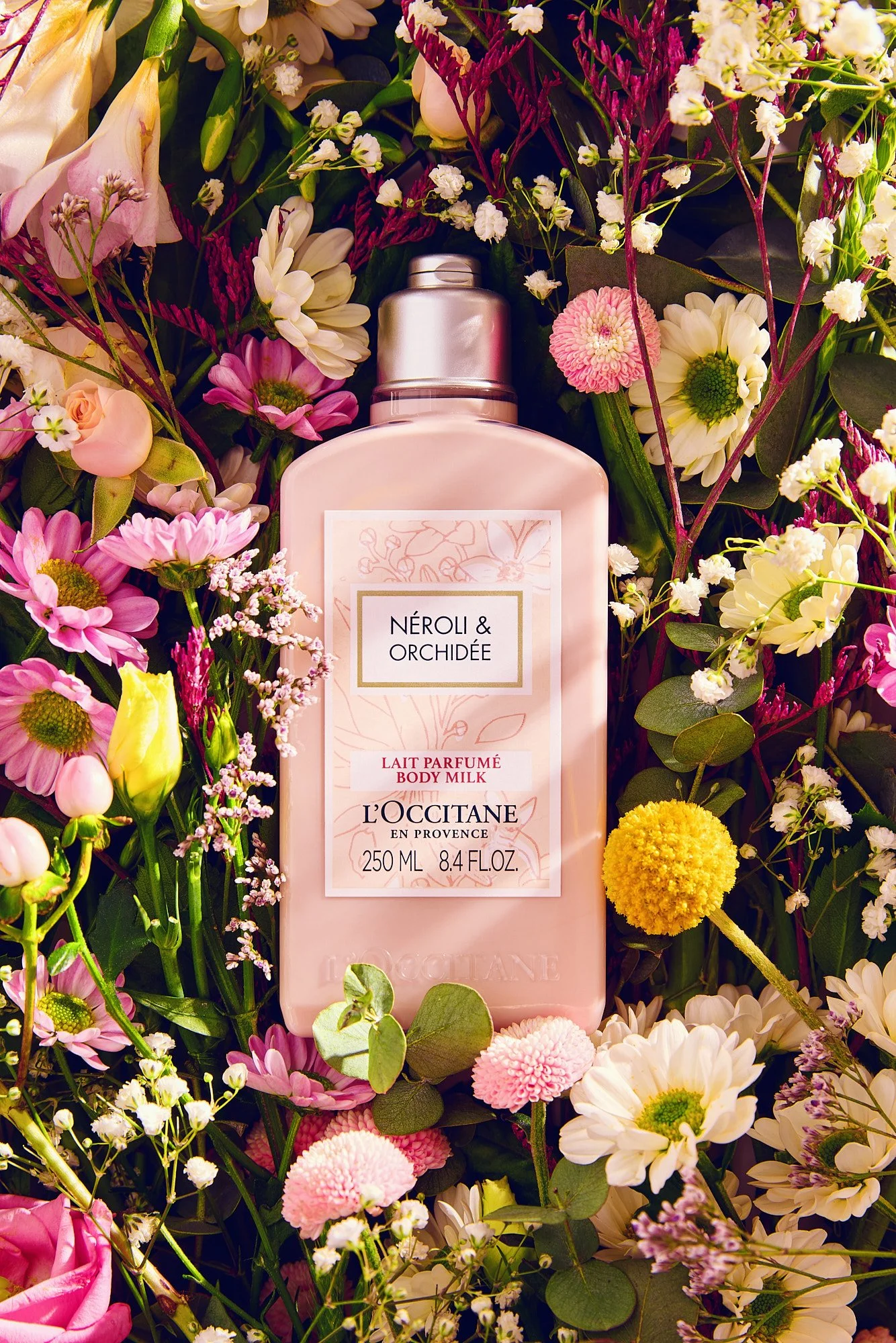 A body milk bottle labeled 'Néroli & Orchidée' surrounded by various pink, white, and yellow flowers and green leaves.