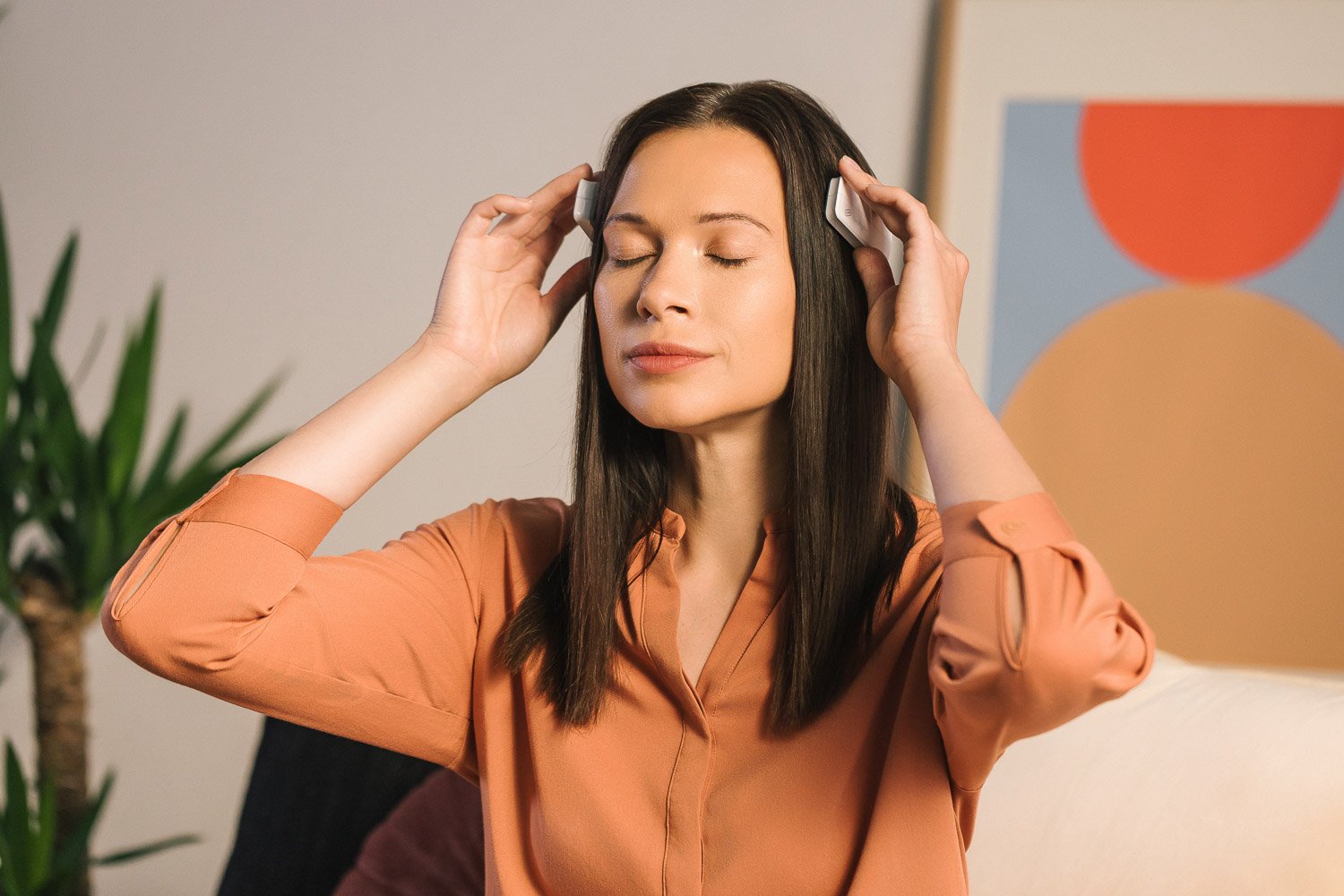 A woman with long dark hair, wearing an orange blouse, is relaxing with eyes closed, listening to music with white headphones in a cozy living room.