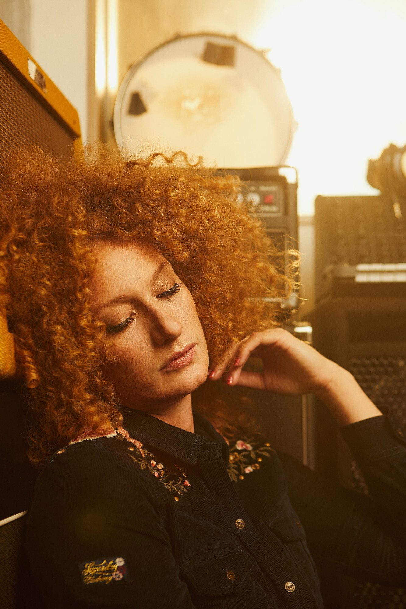 Marina Martensson, A woman with curly red hair, closed eyes, wearing a black embroidered shirt, sitting with her head tilted back and hand touching her neck in a room with musical equipment and a mirror.