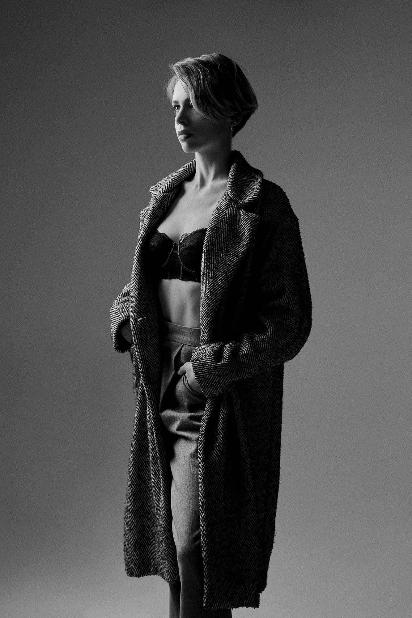 Black and white photo of a woman with a short hairstyle, wearing a long checkered coat, a black top, and light-colored pants, standing against a plain background.