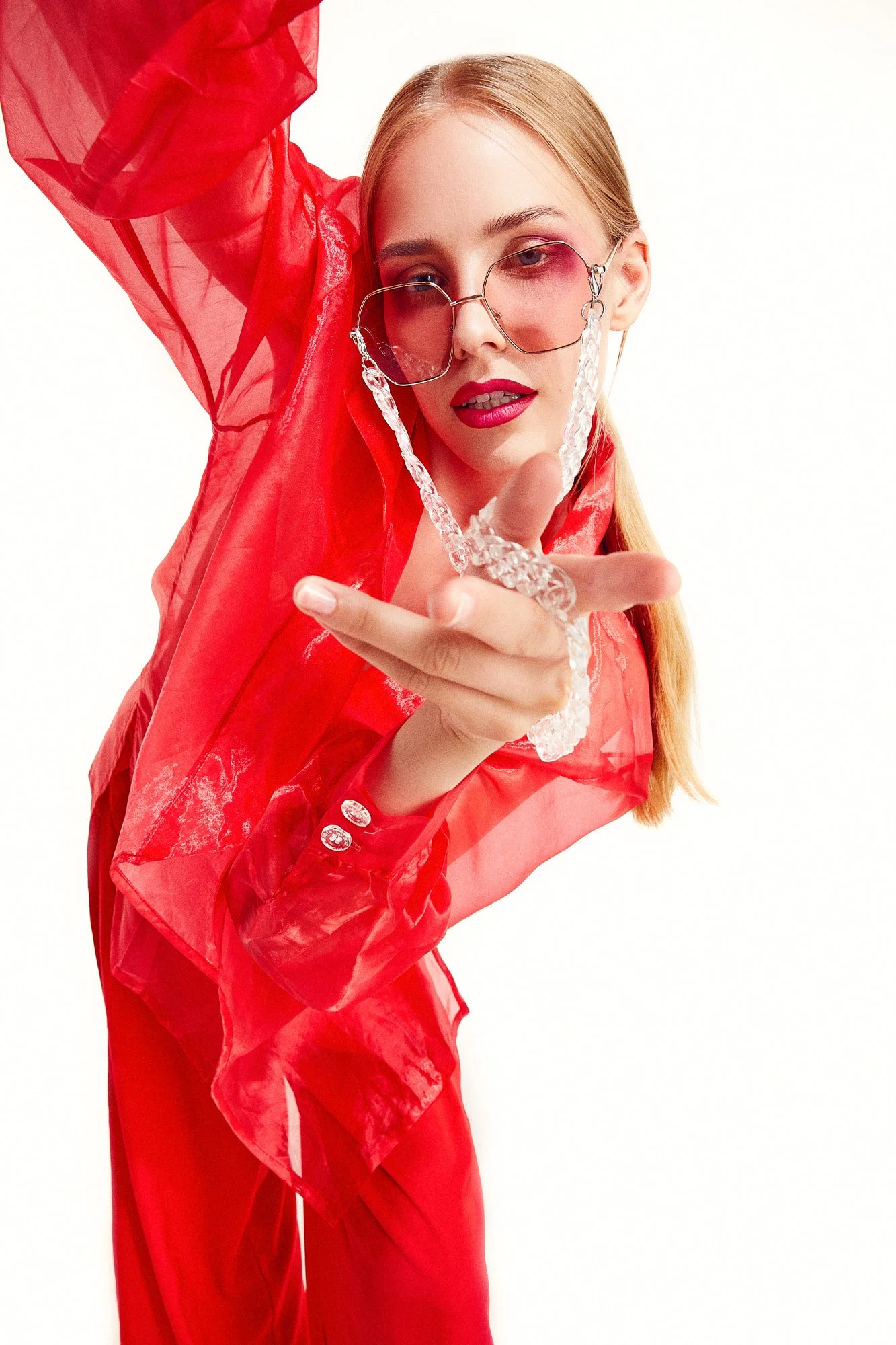 A woman with blonde hair and pink makeup, wearing pink-tinted sunglasses with crystal chain, in a red sheer jacket and red pants, posing against a white background.