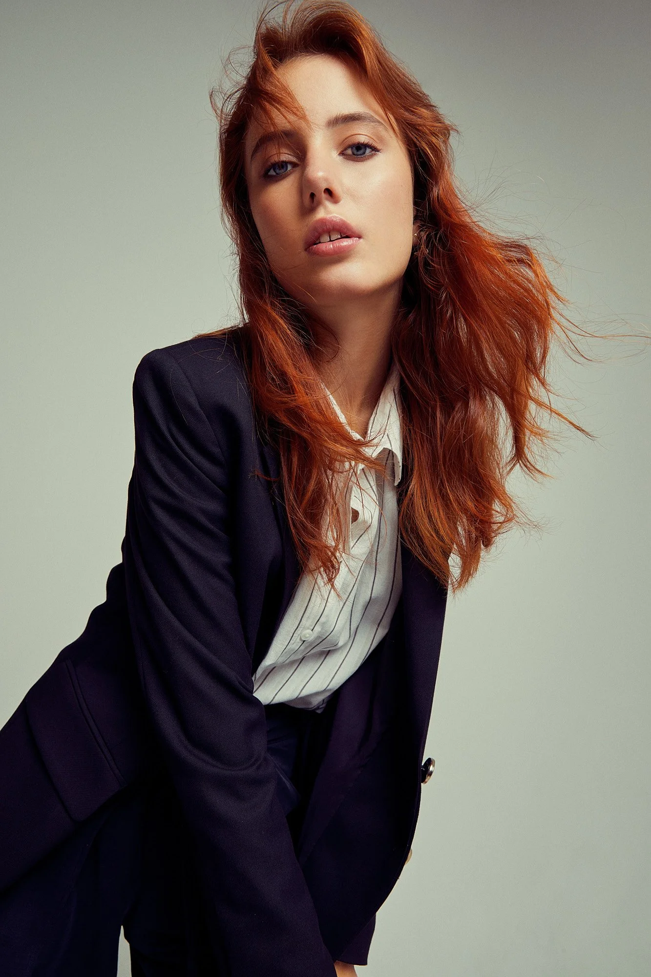 A young woman with red hair and blue eyes wearing a black blazer over a white button-up shirt with brown pinstripes, standing against a plain light grey background.