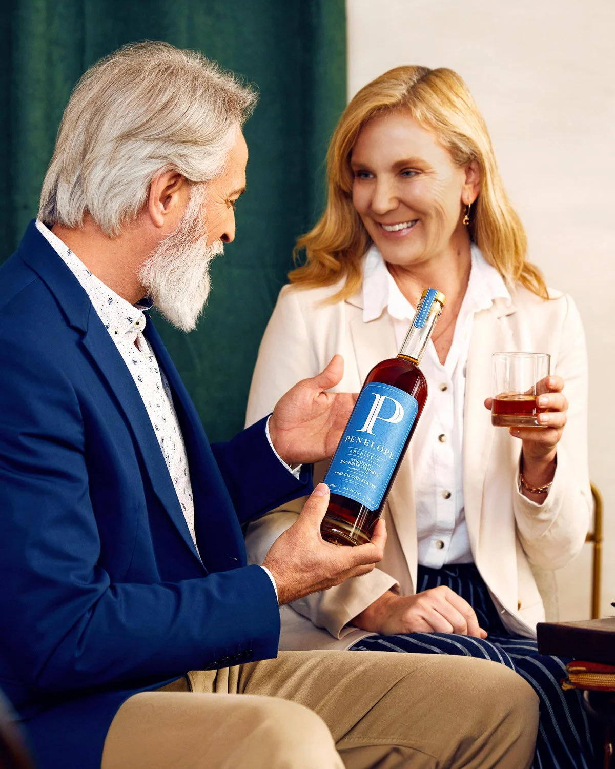 An older man with gray hair and beard in a blue suit presenting a bottle of Penelope bourbon to a woman with blonde hair in a white blazer, who is holding a glass of bourbon.