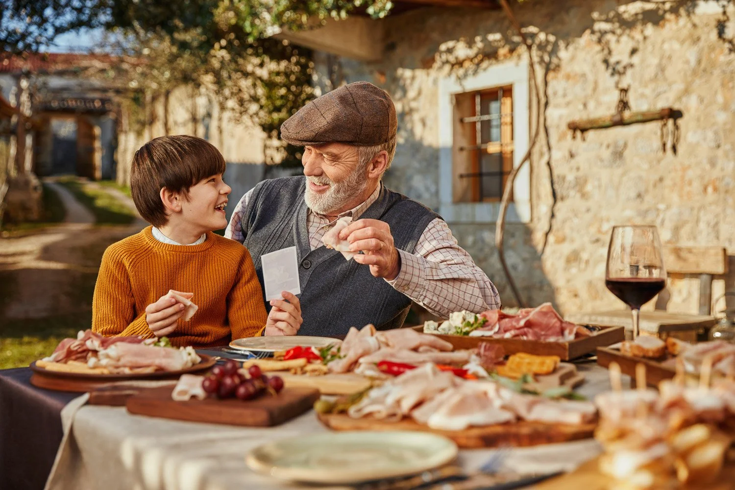 An elderly man and a young boy sharing a joyful moment at an outdoor feast with a rustic stone house in the background. The table is filled with assorted meats, cheese, grapes, and a glass of red wine.