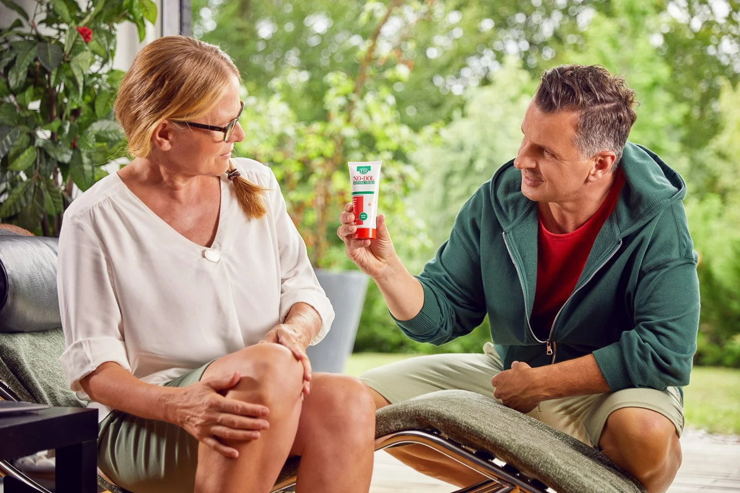 A man is holding a tube of ointment or cream and showing it to an older woman, who is sitting outdoors with greenery in the background. The woman appears to have a knee injury or pain, as she is touching her knee.