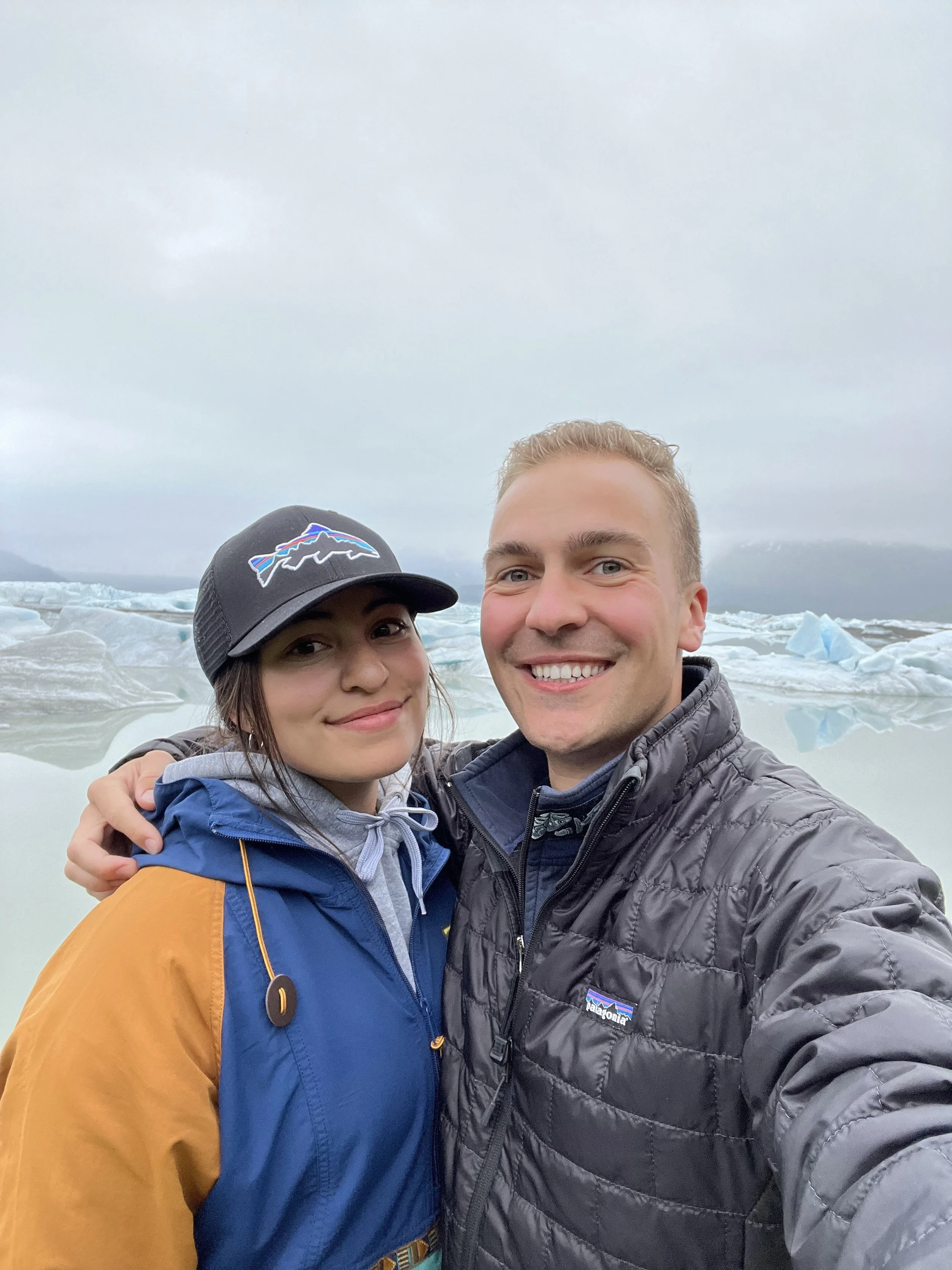 A man and woman smiling and taking a selfie in Cordova, Alaska near a glacier and icebergs, overcast sky, in cold weather