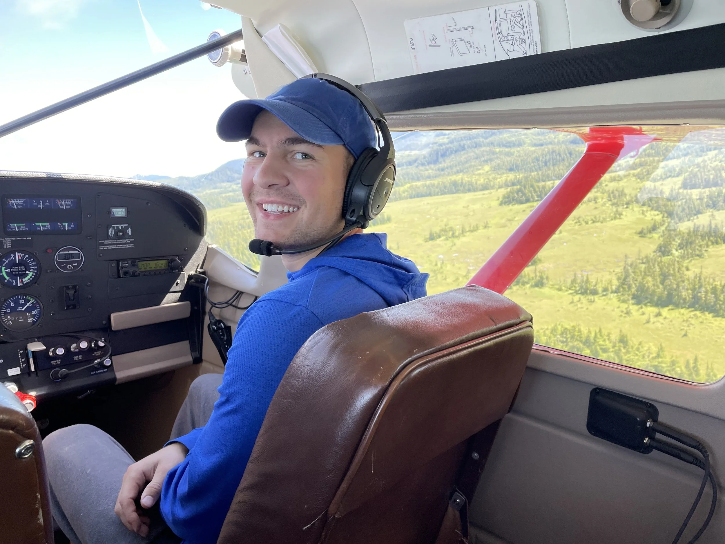 A man in a blue cap and headset sitting in the cockpit of a small airplane, smiling, with open landscape visible through the window.
