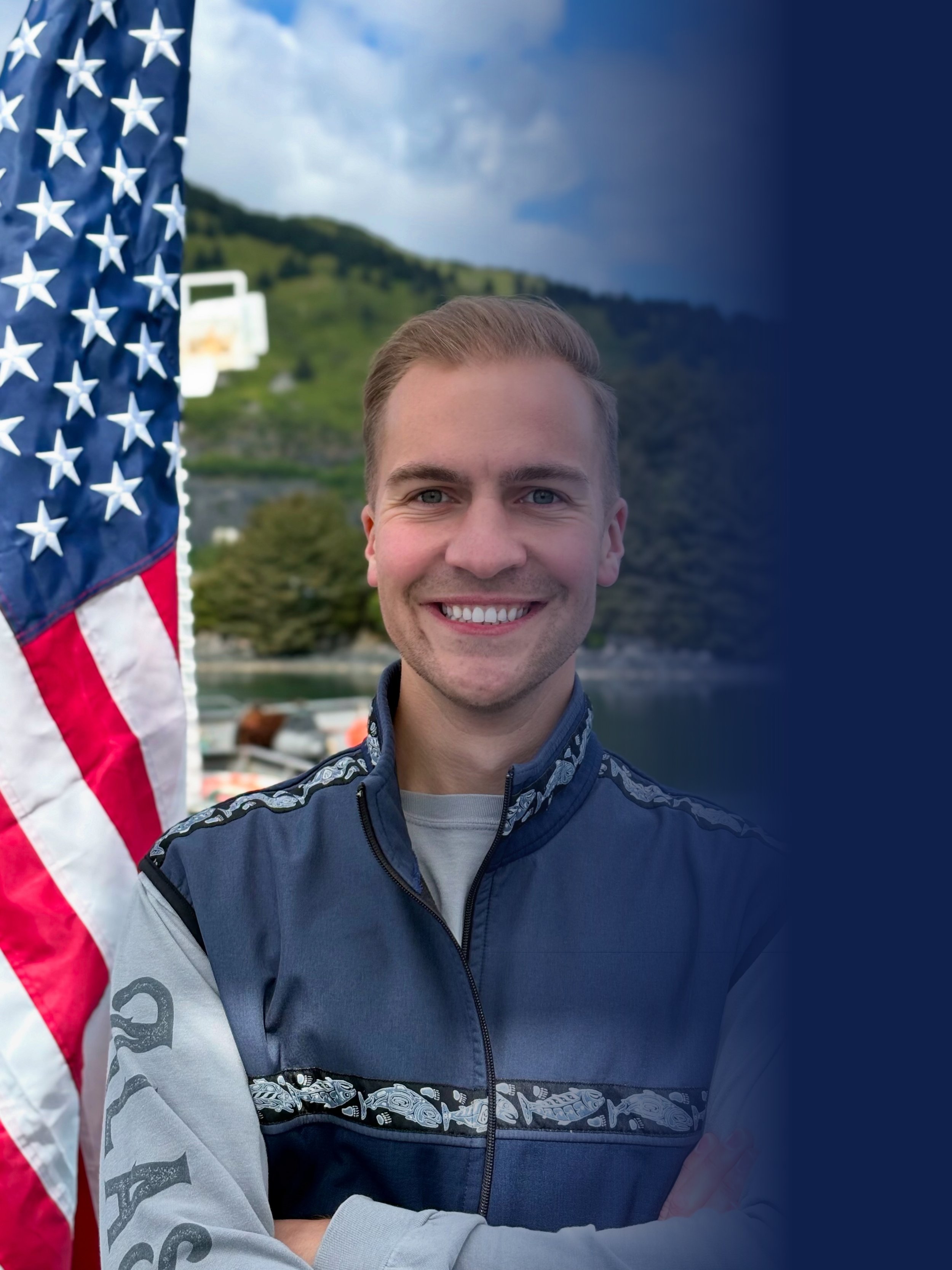 A smiling man with brown hair standing near an American flag outdoors with a Kodiak and harbor in the background.
