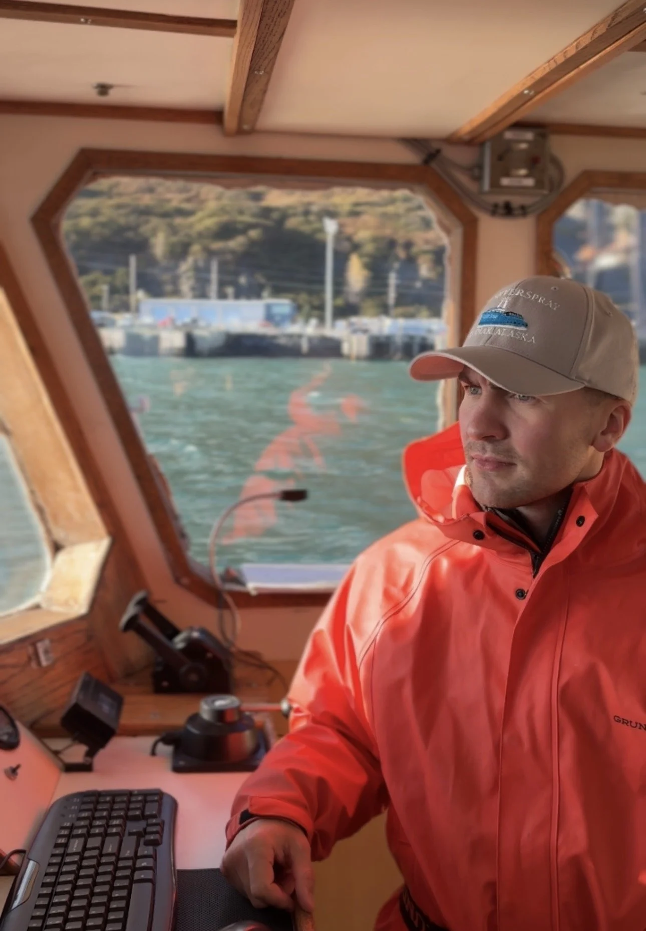 A man wearing a beige cap and red jacket sitting inside a boat's cabin, looking out of the window at a waterfront scene with Kodiak in the background.