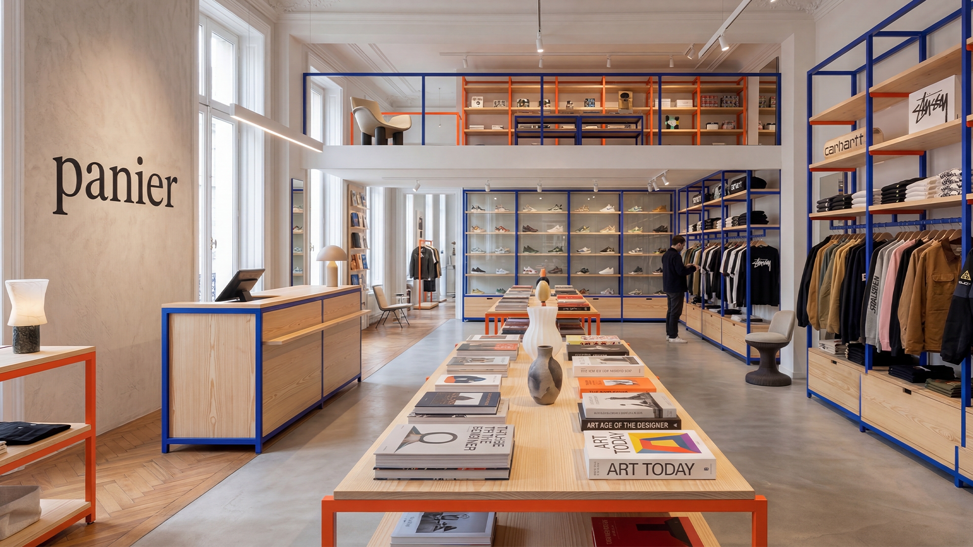 Vitrine d'une boutique de mode avec vêtements, chaussures et accessoires sur des étagères en bois et cadres en bleu et orange, avec un comptoir à la caisse, des tables de livres et des fauteuils, dans un intérieur lumineux avec fenêtres, présentant une ambiance moderne et épurée.