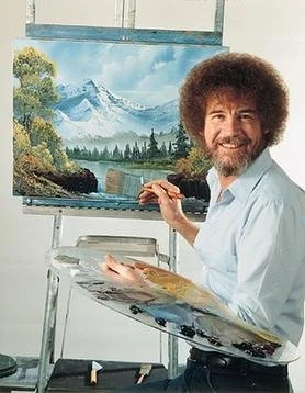 A man with a large curly afro hairstyle and a beard painting a landscape of snow-capped mountains and trees on an easel in a studio.