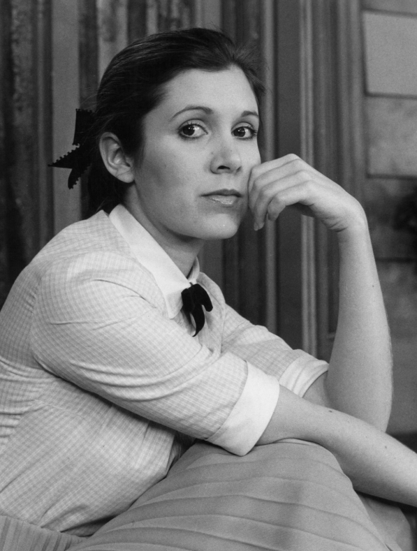 Black and white photo of a woman with dark hair tied back with a bow, sitting at a table with her hand resting on her chin, looking at the camera with a contemplative expression.