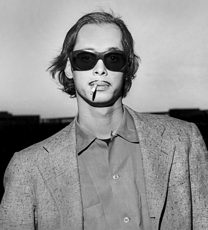 A person with shoulder-length hair wearing sunglasses, a cigarette in their mouth, a collared shirt, and a blazer, standing outdoors with a blurred background.