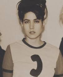 Young woman with dark hair styled in an updo, wearing a beige T-shirt with a black collar and a large black number '3' on the front.