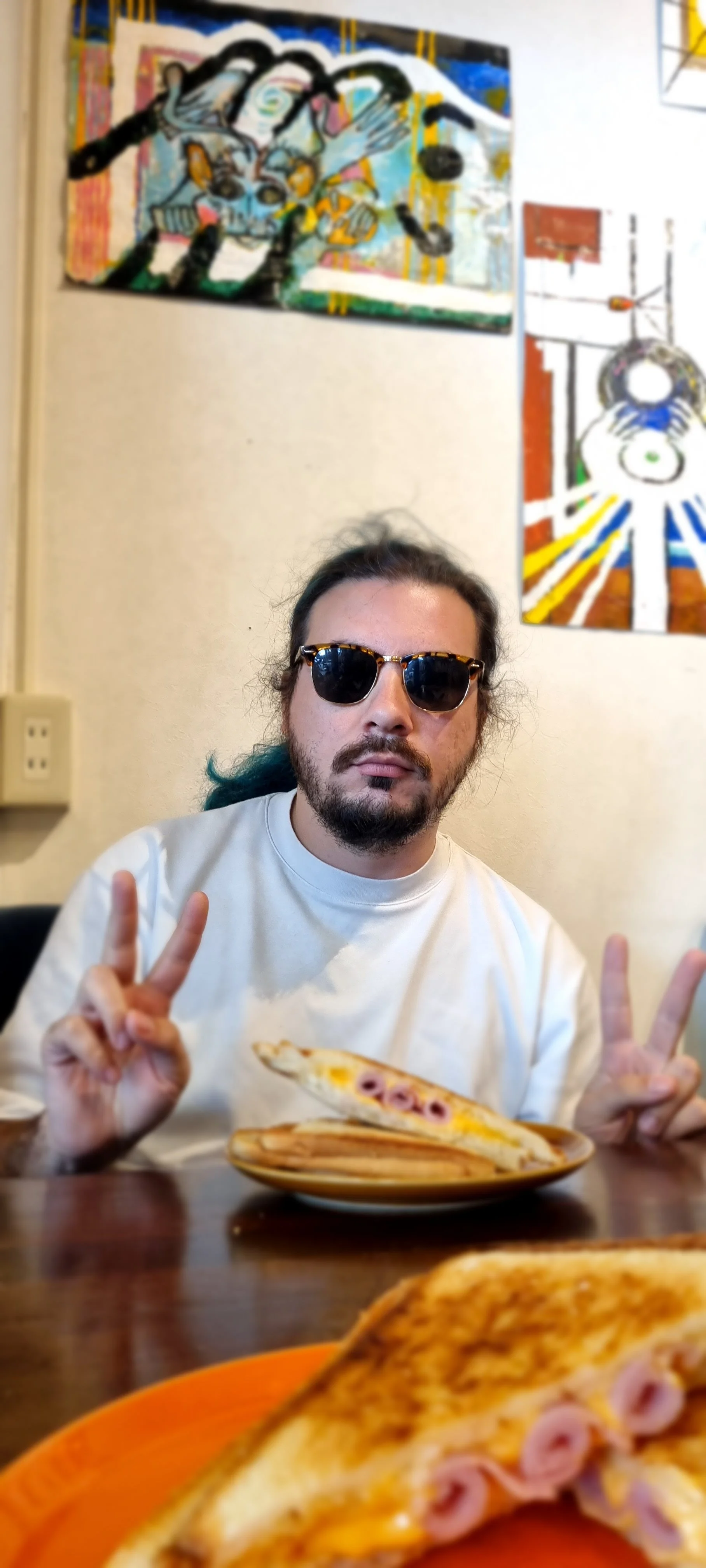 A man with long dark hair, sunglasses, and a beard making peace signs with both hands at a table with a slice of pizza on a plate. There are colorful abstract paintings on the wall behind him.