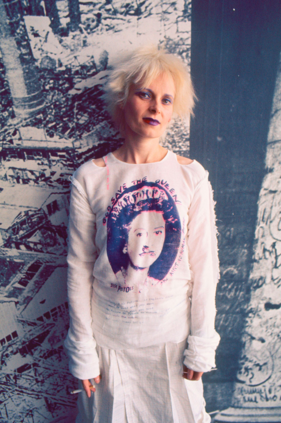 A woman with bleached, tousled hair and dark lipstick stands in front of a black-and-white mural of city buildings and snowy trees.