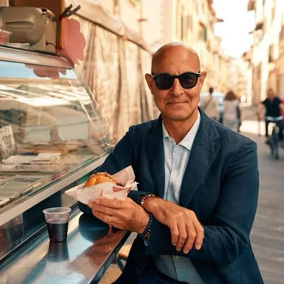 A man wearing sunglasses and a suit jacket sits at a street food cart, holding a sandwich wrapped in paper. There is a drink on the counter, and the background shows a city street with buildings and pedestrians.