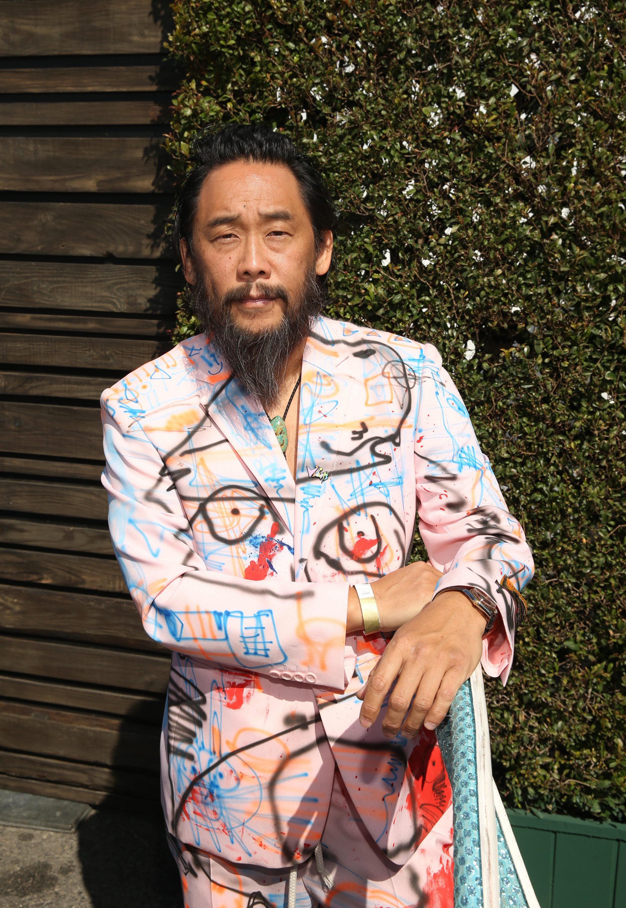 A man with dark hair and a beard wearing a jacket covered in colorful abstract graffiti, standing outdoors in front of a wooden fence and green shrubbery.